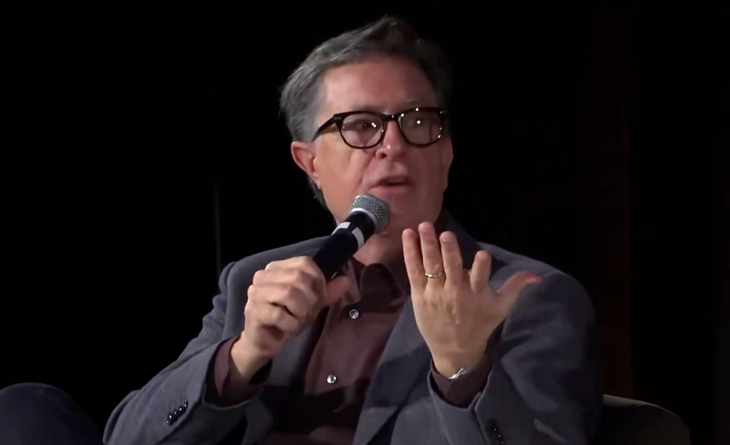 En person i briller og dress snakker i en mikrofon under et arrangement