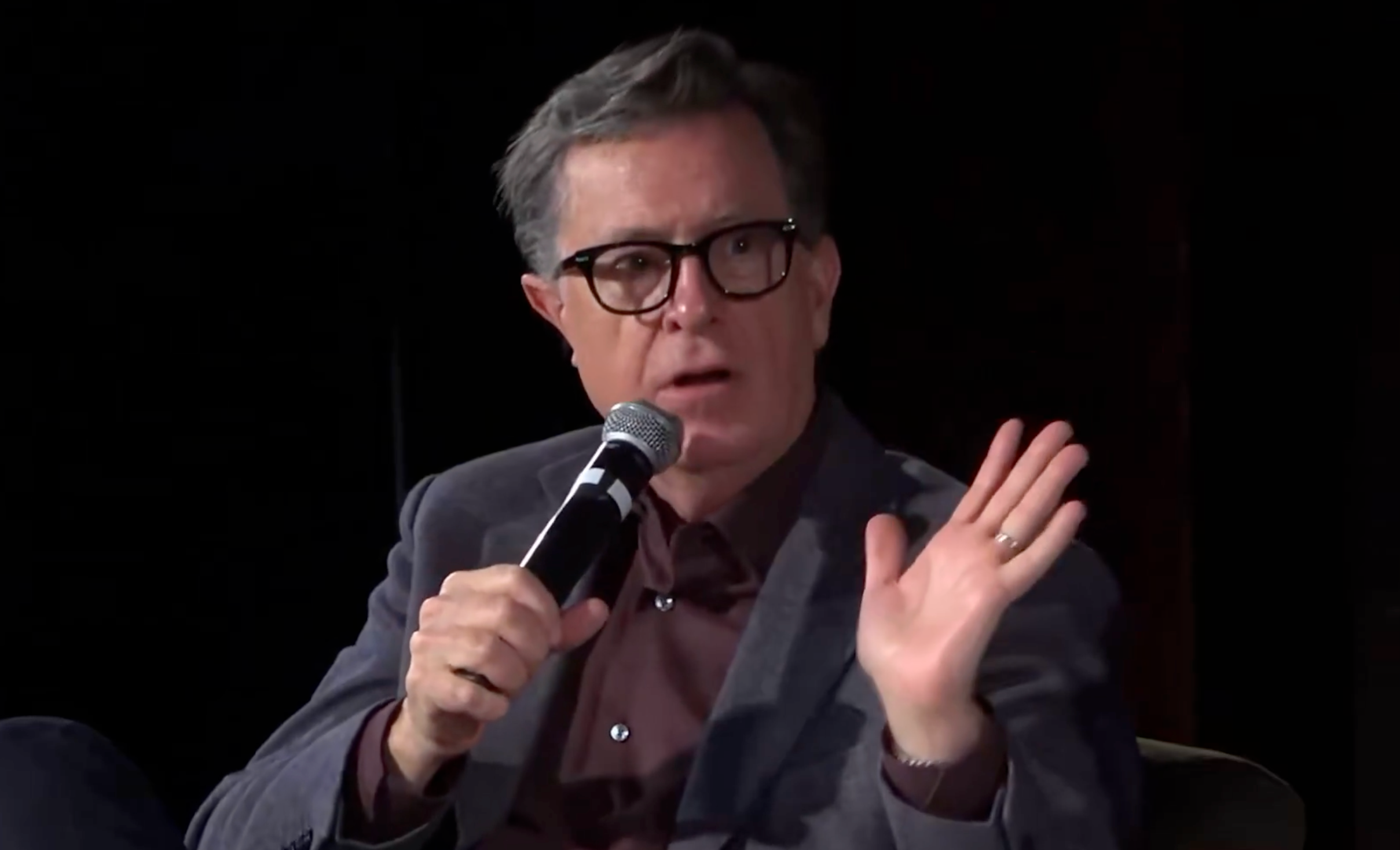 A idiosyncratic   wearing glasses, successful  a suit, speaks into a microphone portion    gesturing with their manus  during an event