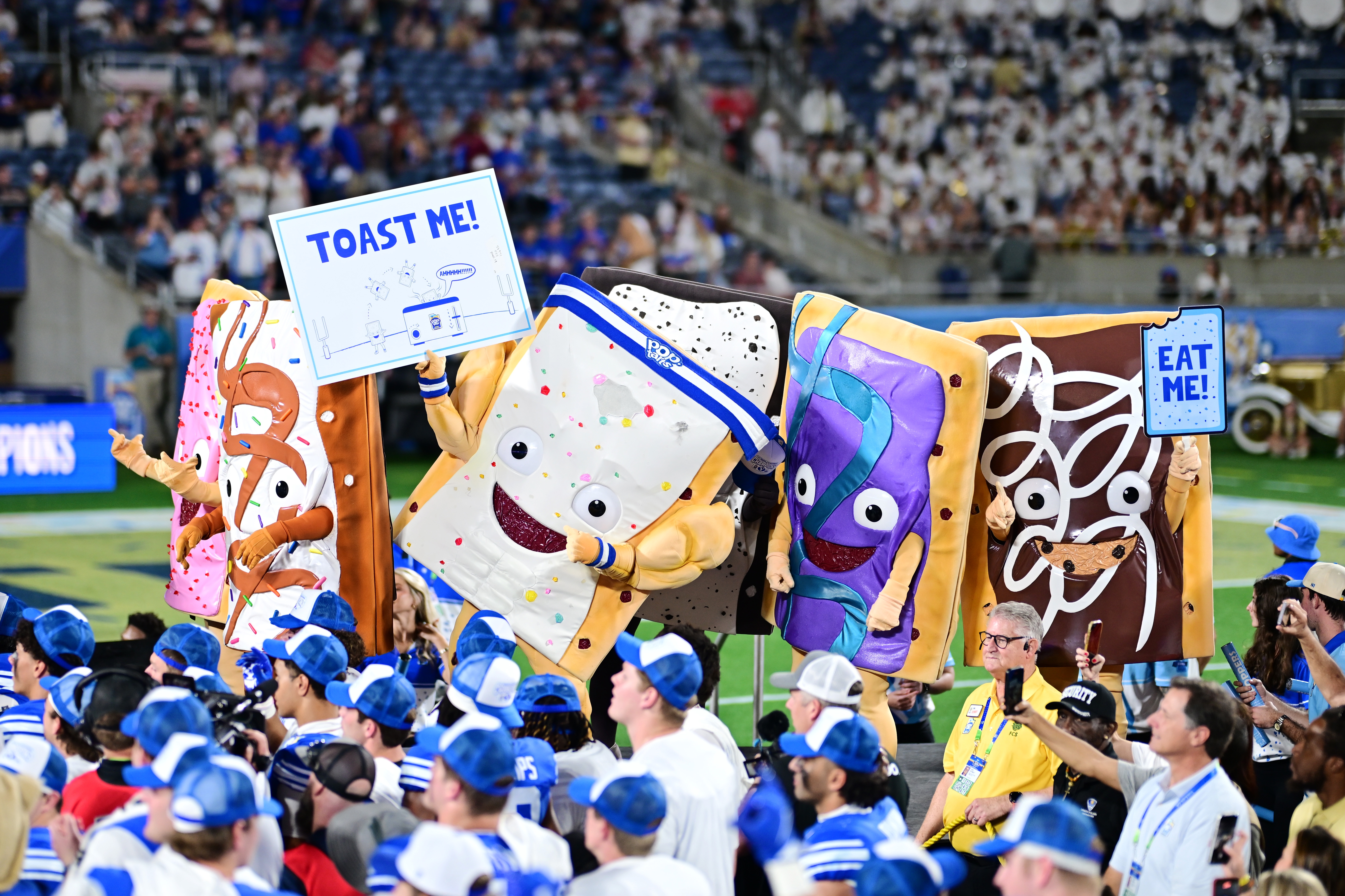 Characters dressed arsenic  playful Pop-Tart mascots clasp  signs speechmaking  "TOAST ME!" and "EAT ME!" astatine  a sports event, surrounded by cheering fans