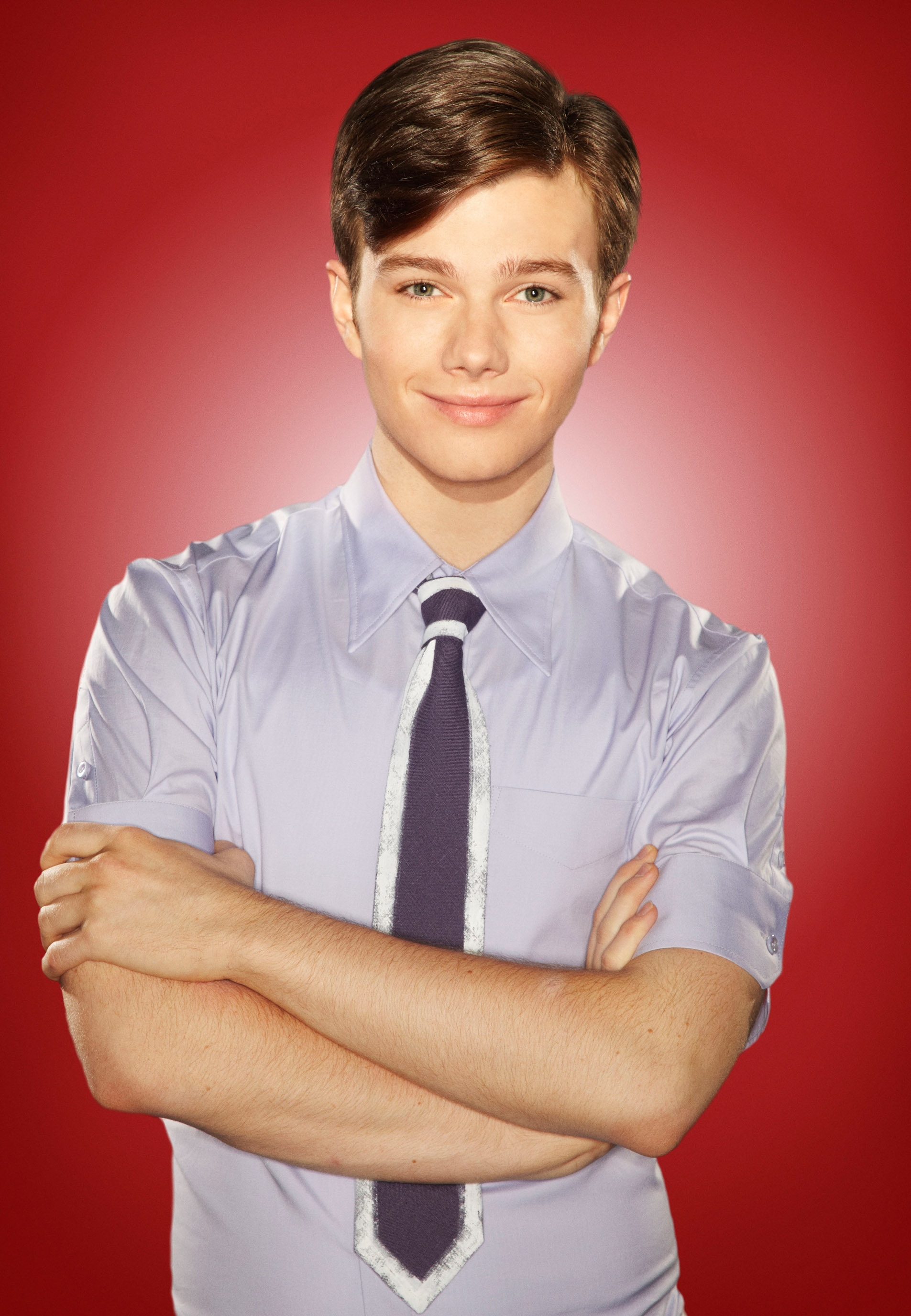 Person with neatly styled hair, wearing a airy  shirt, tie, and folded arms, lasting  against a plain backdrop