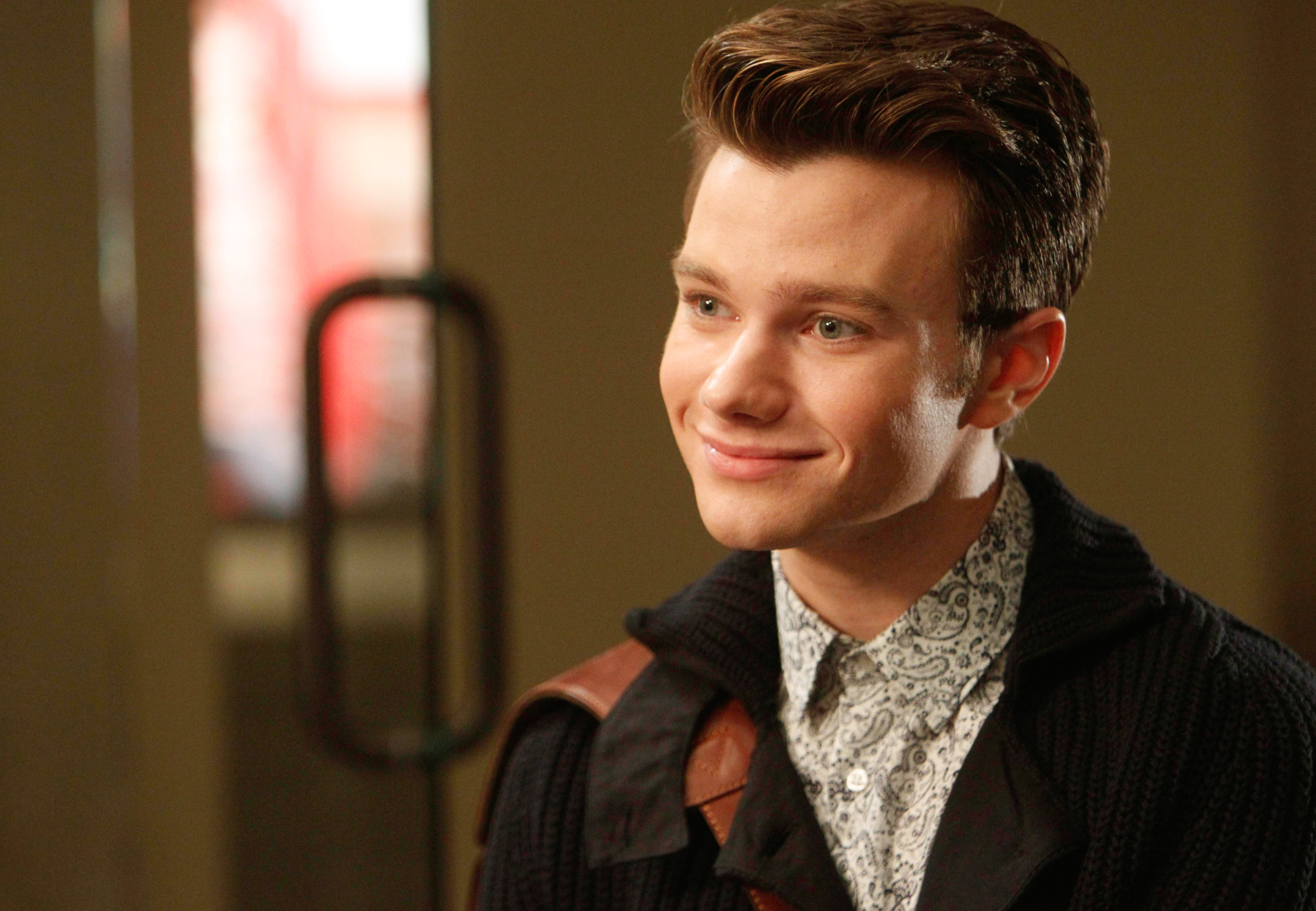 Chris Colfer smiling, wearing a patterned garment  and acheronian  cardigan, looking somewhat  to the broadside  indoors