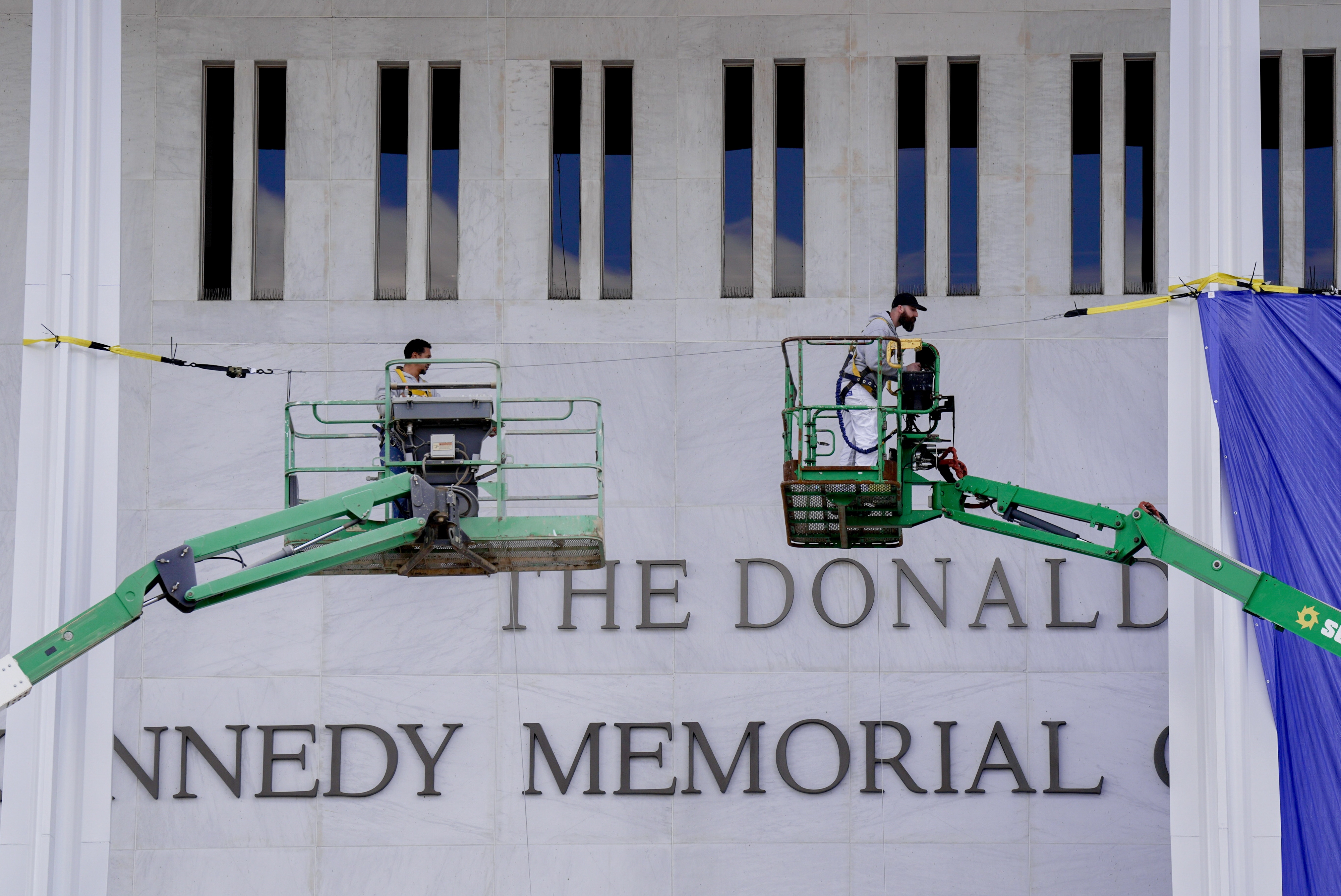 Workers on lifts cover the Kennedy Center sign with a large fabric, partially hiding the text on the building