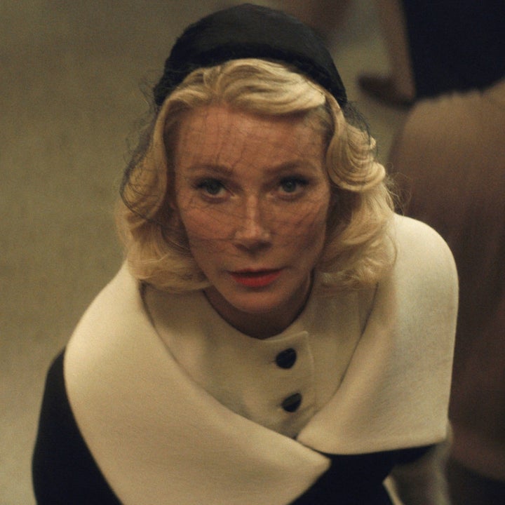 Person in vintage fashion, wearing a veiled hat and a white fur-trimmed coat, looks up amid a crowd. 1950s-style scene