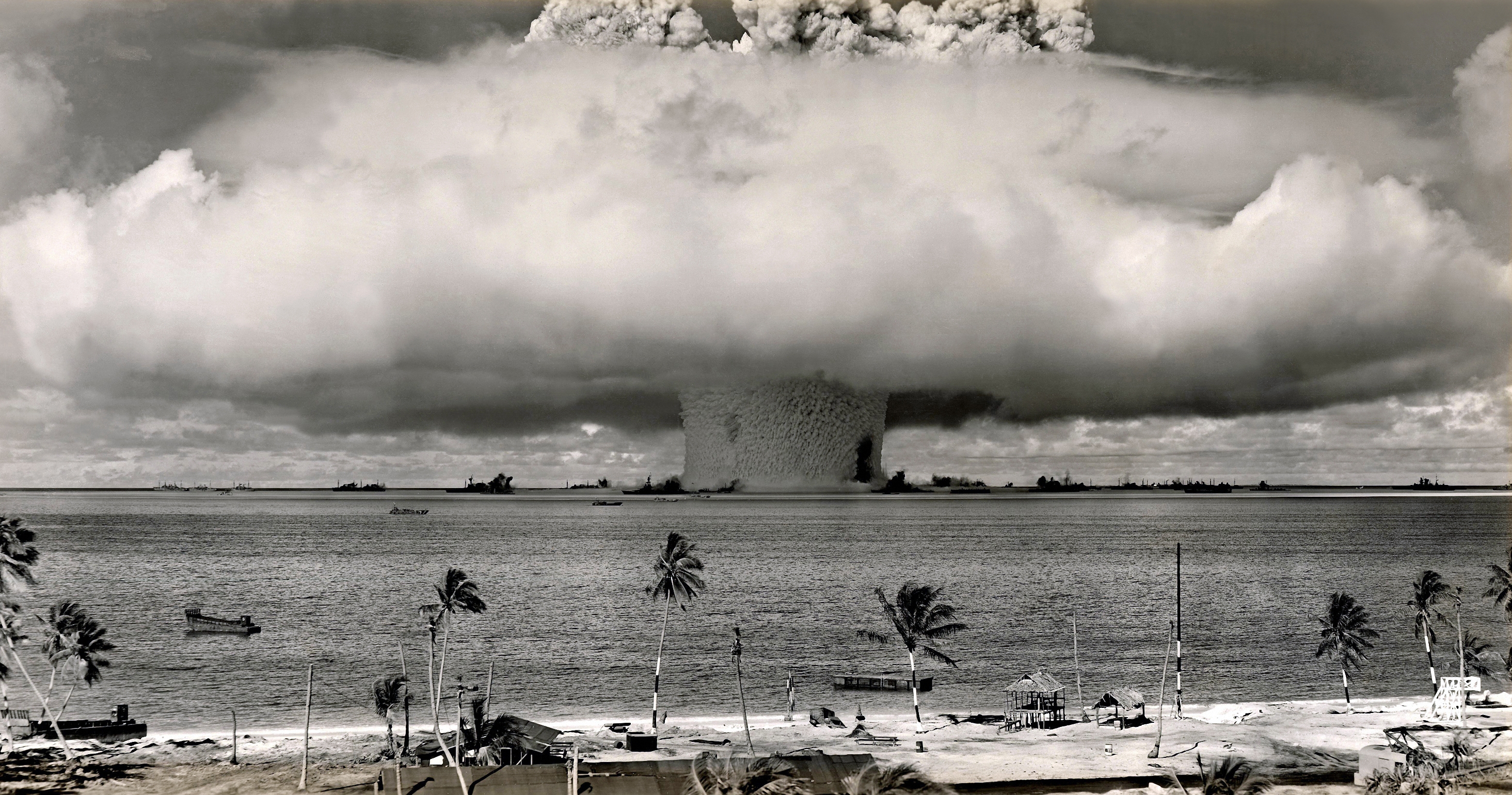 A historical black-and-white photo of a nuclear explosion with a large mushroom cloud rising over the ocean, viewed from a shoreline with palm trees