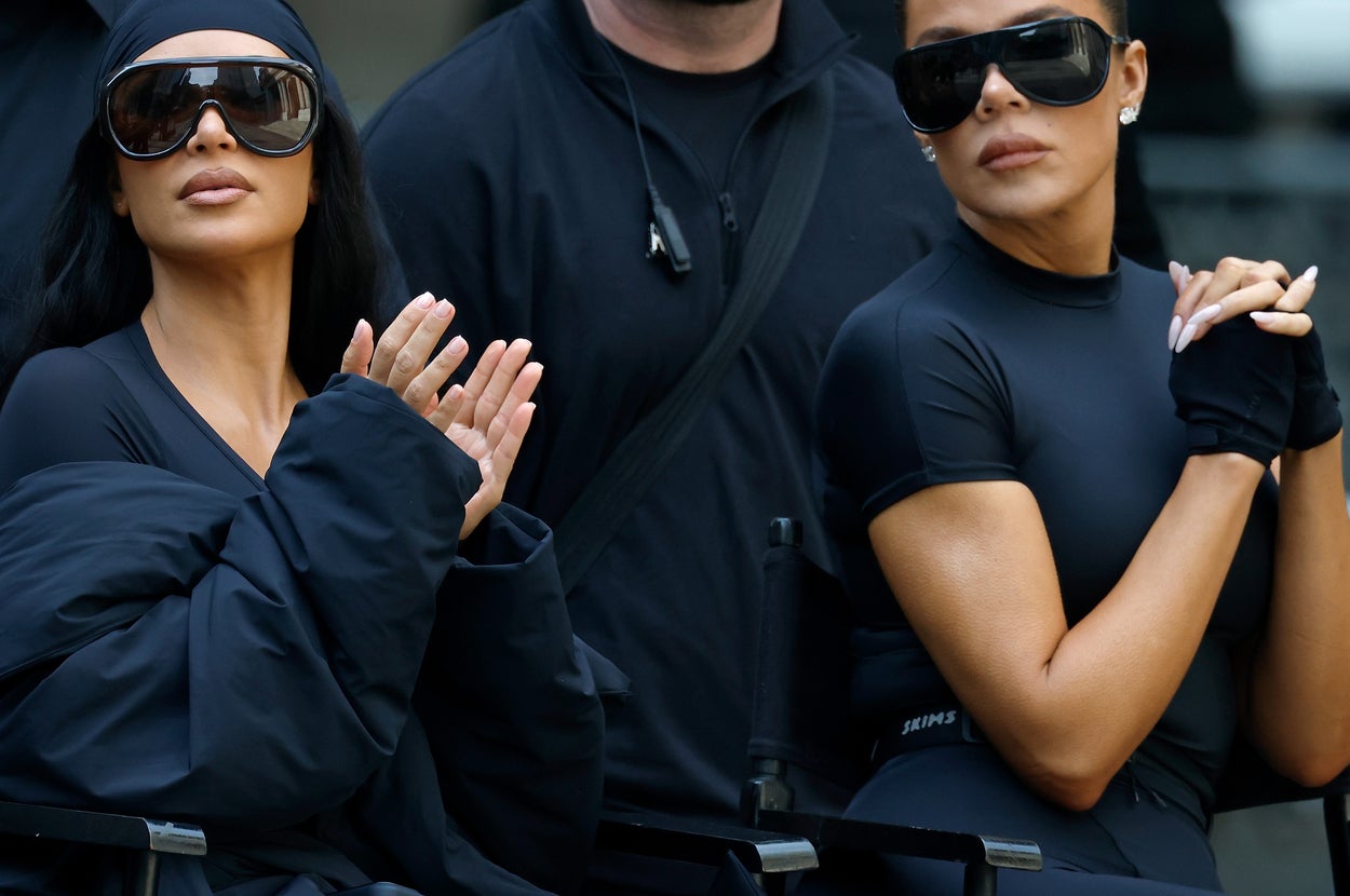 Kim and Khloe Kardashian in sleek attire and oversized sunglasses attend an outdoor event, sitting next to each other and appearing focused