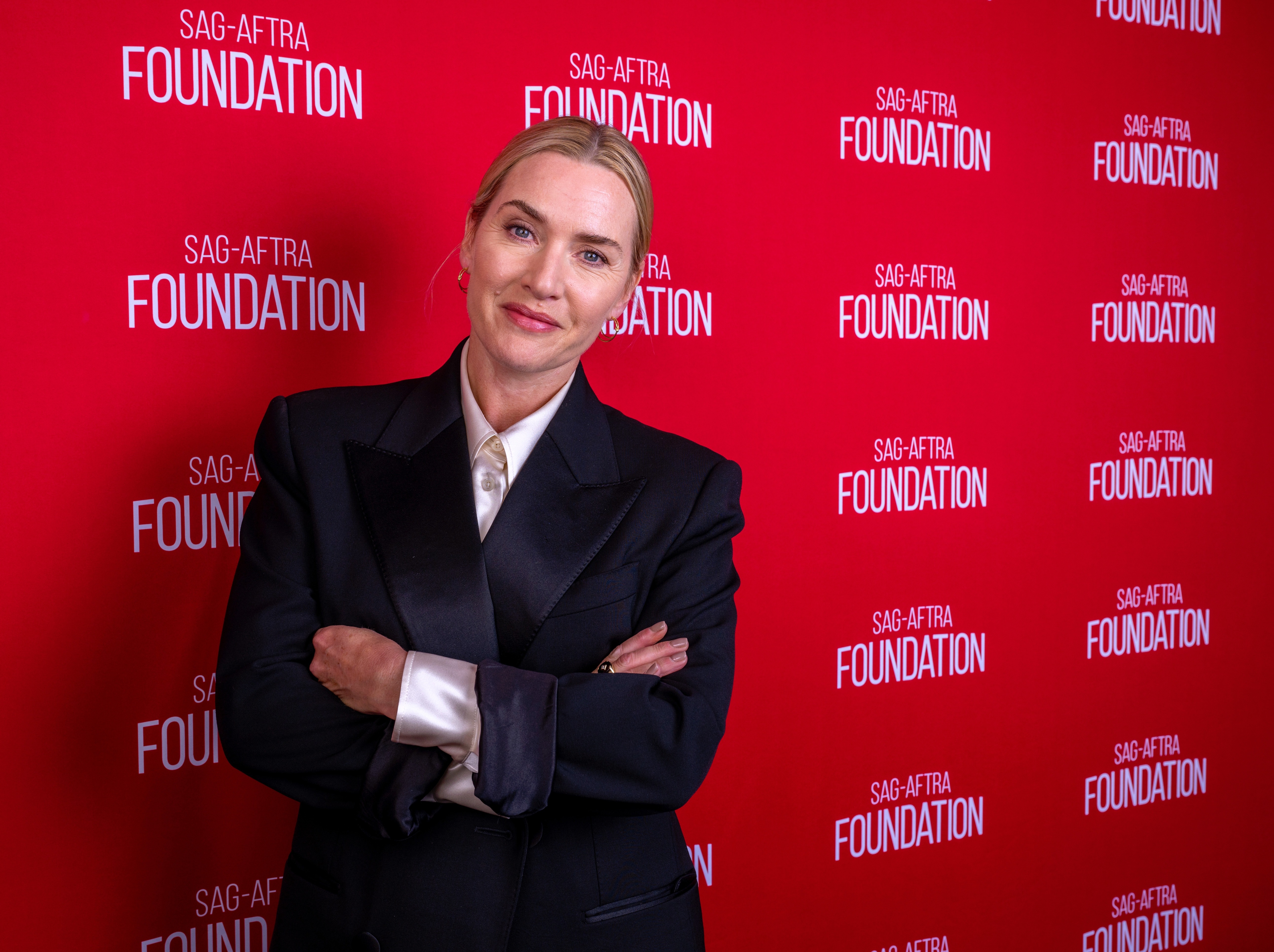 Kate Winslet successful  a stylish suit   stands with arms crossed astatine  an lawsuit   with a SAG-AFTRA Foundation backdrop