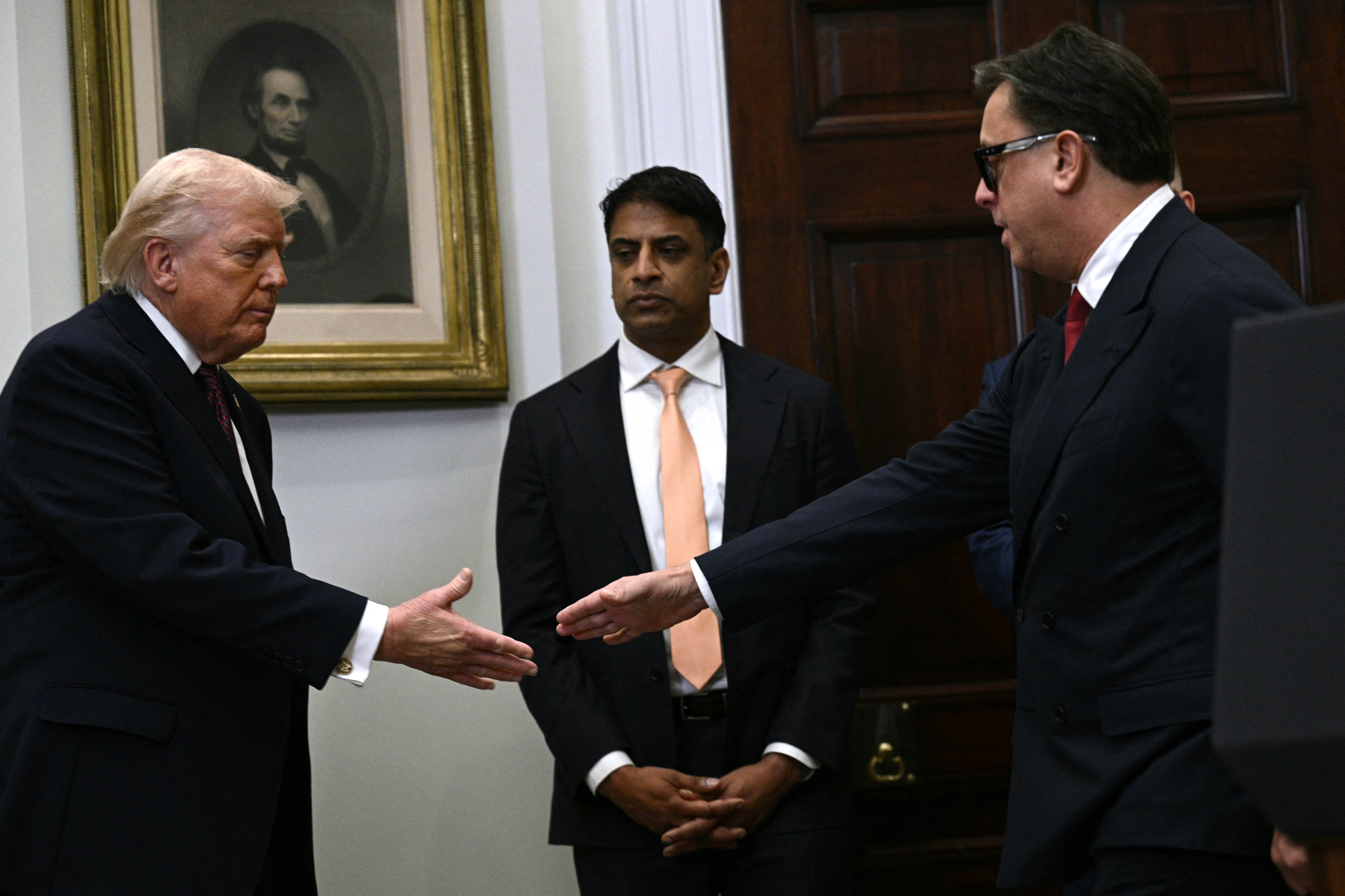 En mann i dress strekker seg for å håndhilse på en annen mann i en dress mens en tredje mann ser på i en formell setting