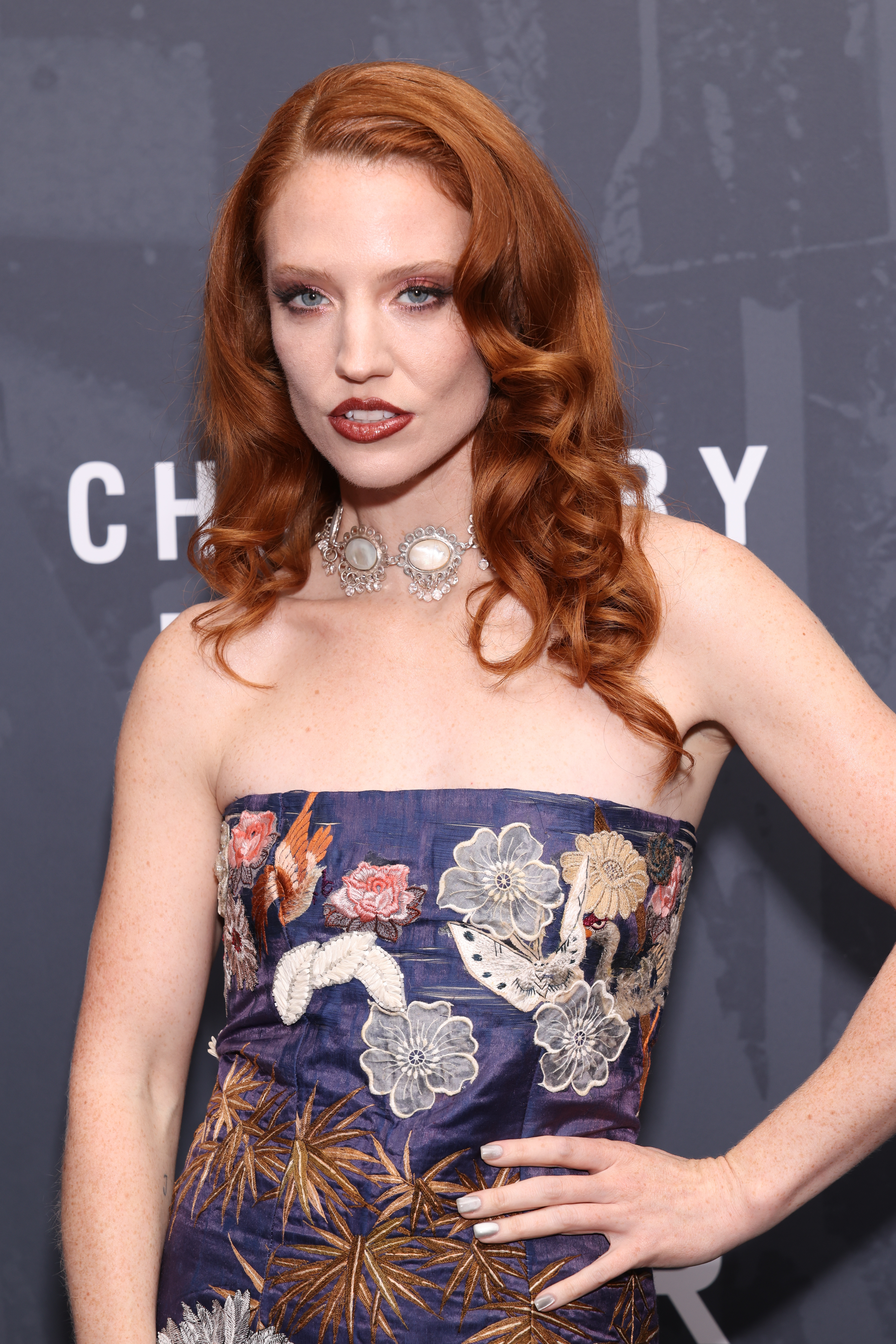 Person med langt bølget hår i en stroppeløs blomsterkjole poserer på et teppe på et offentlig arrangement