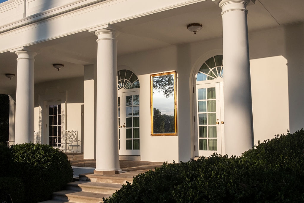 A framed reflector is humorously placed betwixt the doors connected a columned porch, reflecting a histrion successful the background