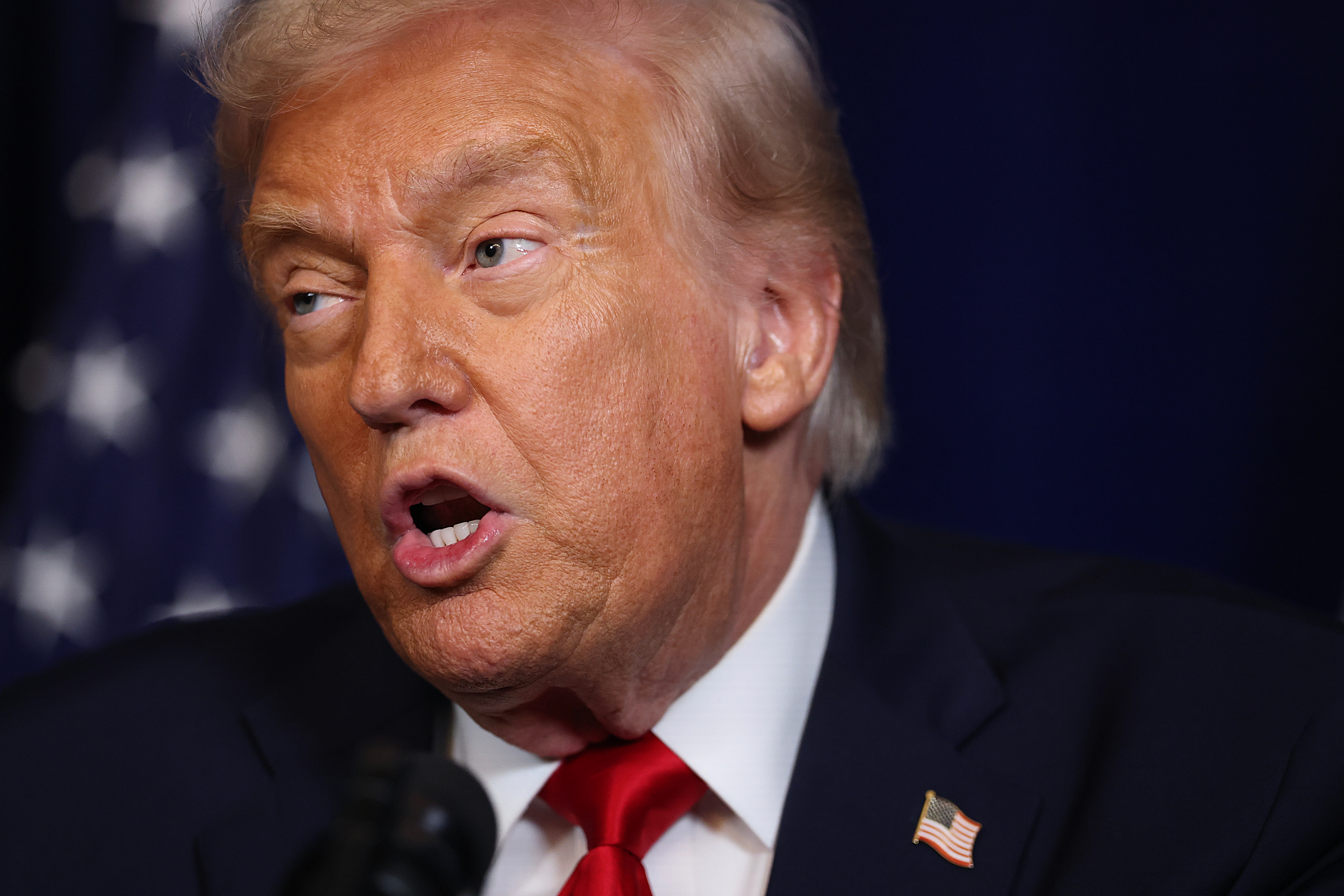 En person i dress og slips snakker på et arrangement med et amerikansk flagg synlig i bakgrunnen