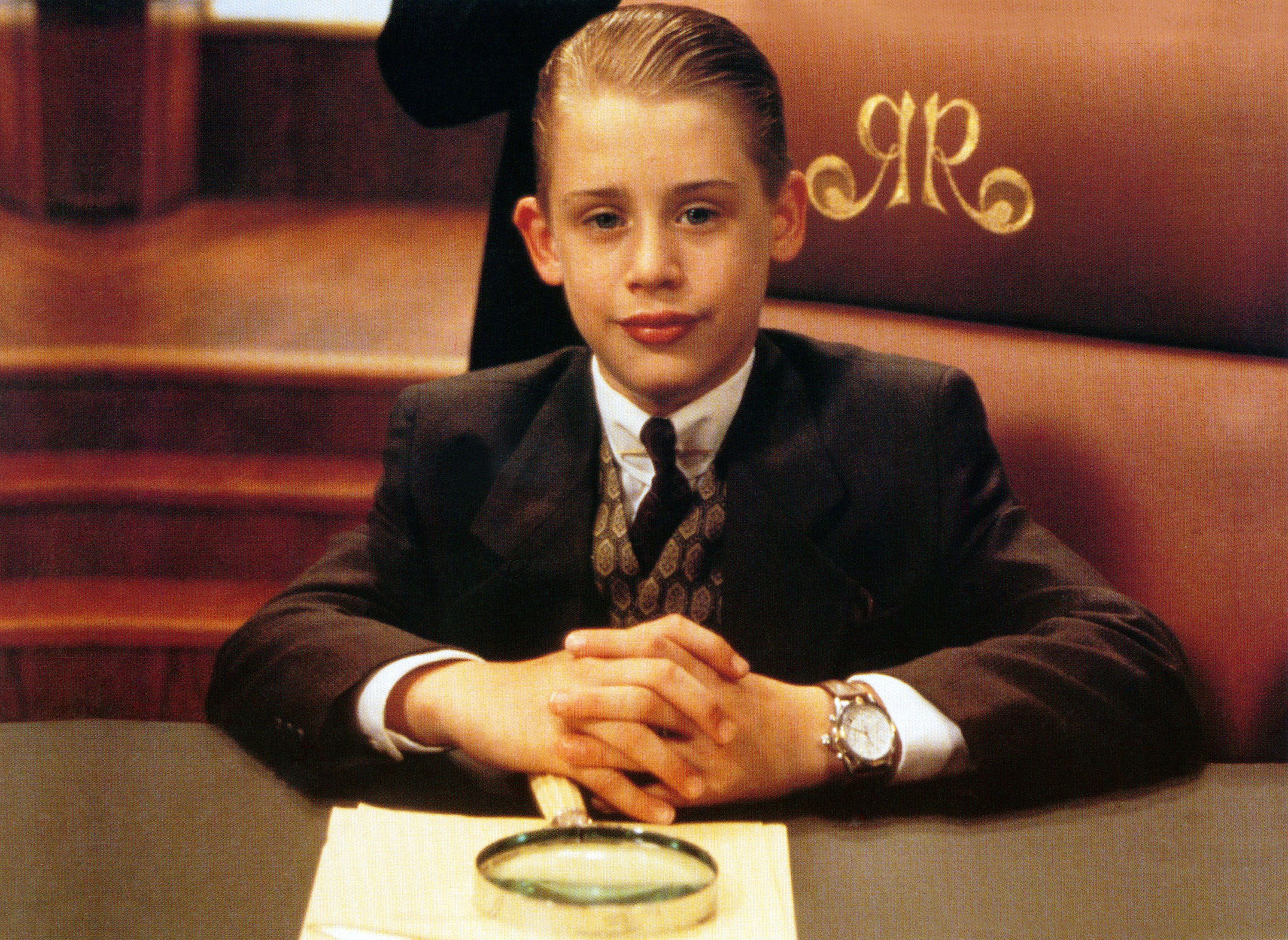 Macaulay Culkin successful  a suit   and necktie  sits confidently astatine  a table  with a monogrammed chair. A magnifying solid  is connected  the array  earlier  him