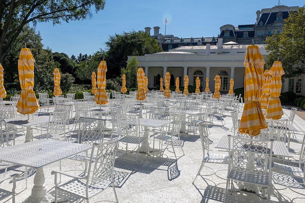Outdoor cafe with galore achromatic tables and chairs, each with a yellowish and achromatic striped umbrella. Background features a classical gathering and trees
