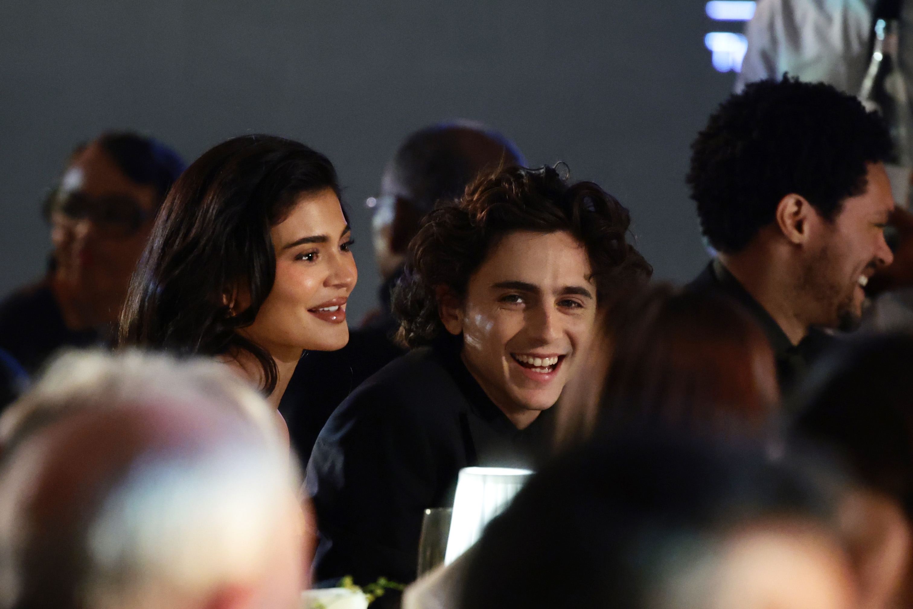 Kylie Jenner and Timothee Chalamet smiling and conversing astatine  a ceremonial  event, seated among others. They look  to beryllium  enjoying the occasion