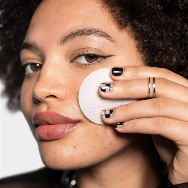 Person applying skincare merchandise with a circular pad to their cheek. They person rings connected their digit and manicured nails with a geometric achromatic design
