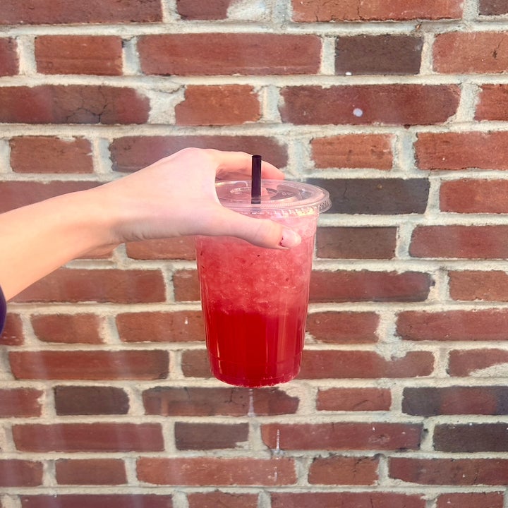 Someone holding a vibrant iced drink in a plastic to-go cup against a brick wall