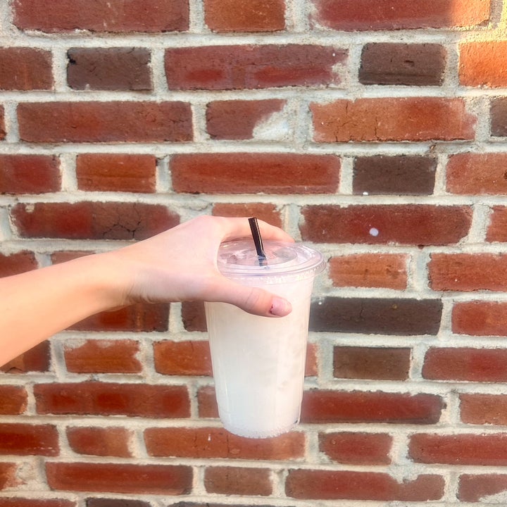 a hand holding up a plastic to-go cup with a lid and straw in front of a brick wall