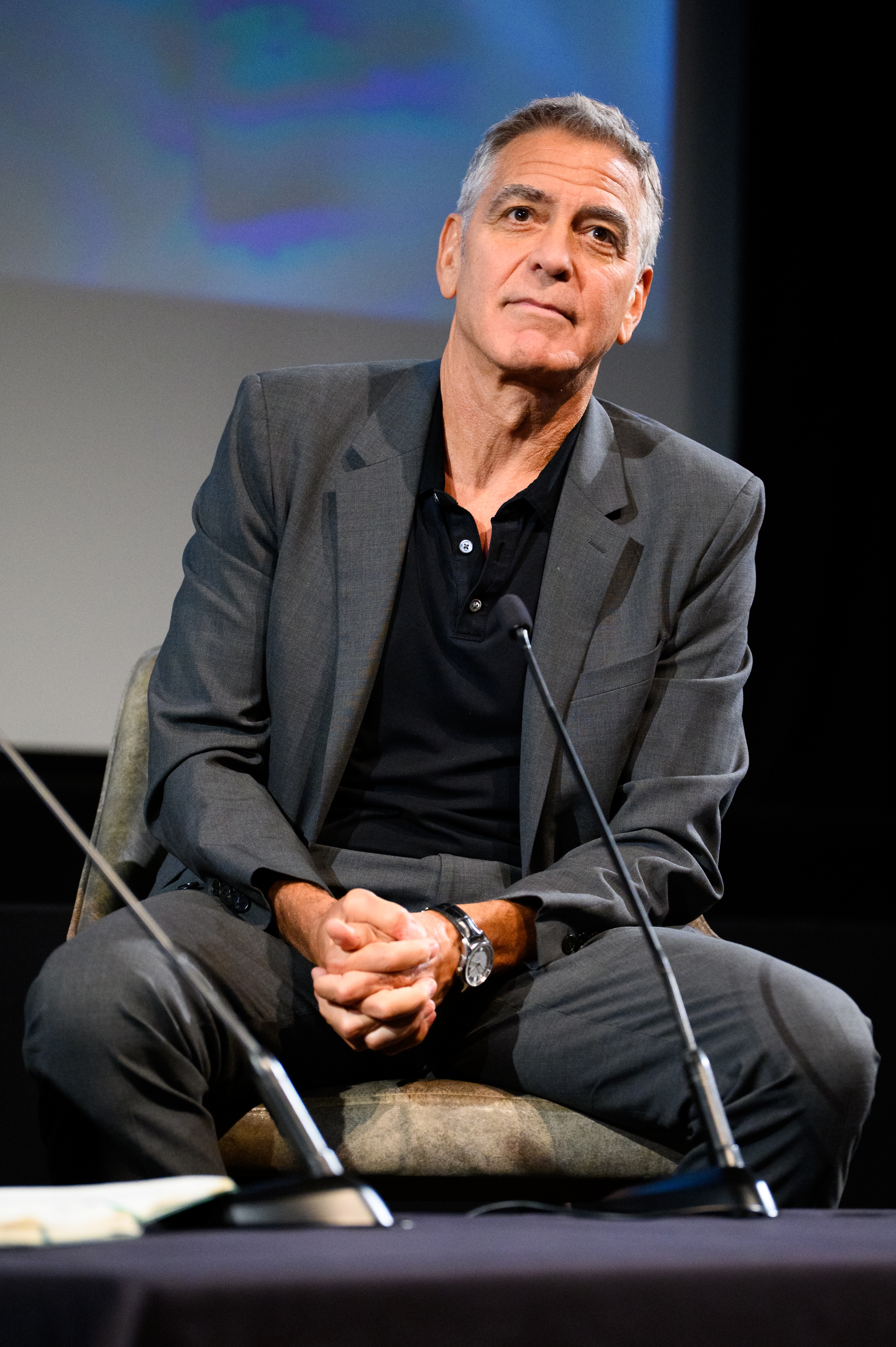 Person seated, wearing a stylish suit   with an open-collar shirt, astatine  a speaking lawsuit   connected  stage