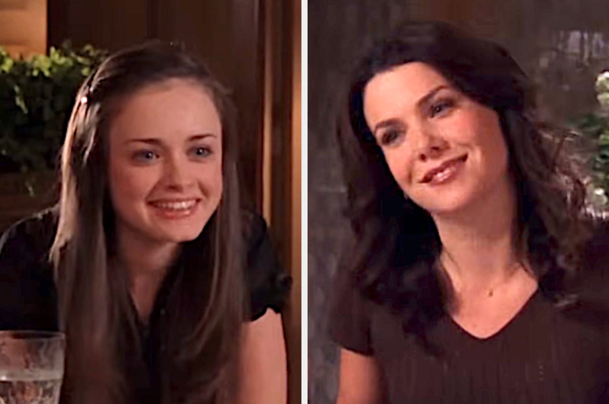 Two women smiling while seated at a table, one younger with long hair and the other older with wavy hair, in a casual setting