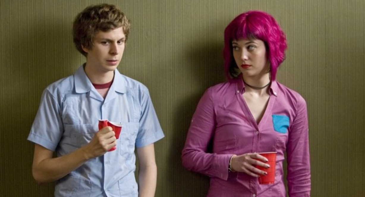 To personer holder røde kopper, stående mot en vegg. Personen til venstre har på seg en fritidsskjorte; personen til høyre har slående rosa hår og en fargerik topp