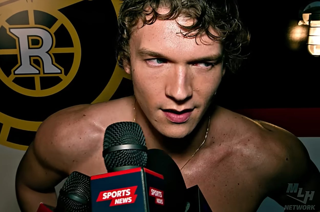 Ilya shirtless speaking into microphones, sports logo behind him