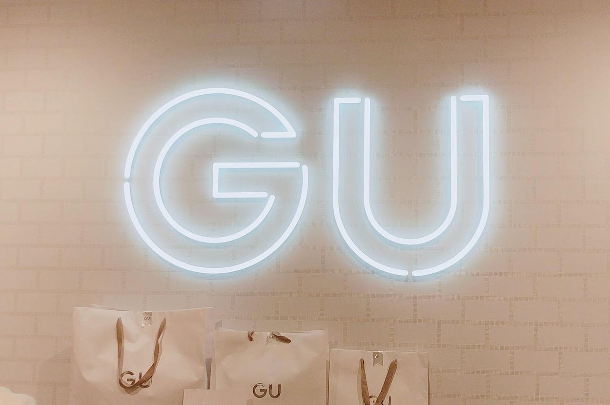 Large illuminated "GU" logo on wall above several shopping bags marked "GU." Sign indicates gift wrapping at an additional cost