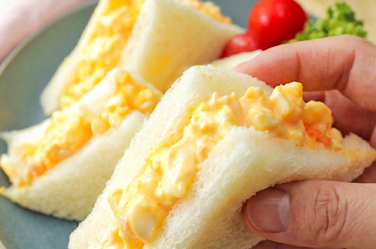 卵サンドイッチを手に持った状態で、添え物にトマトと緑の野菜がある。