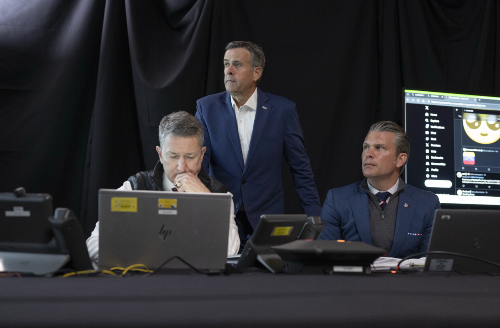 Tre personer i dress sitter og står ved et bord med bærbare datamaskiner og kommunikasjonsenheter, og virker fokuserte og engasjerte i en oppgave