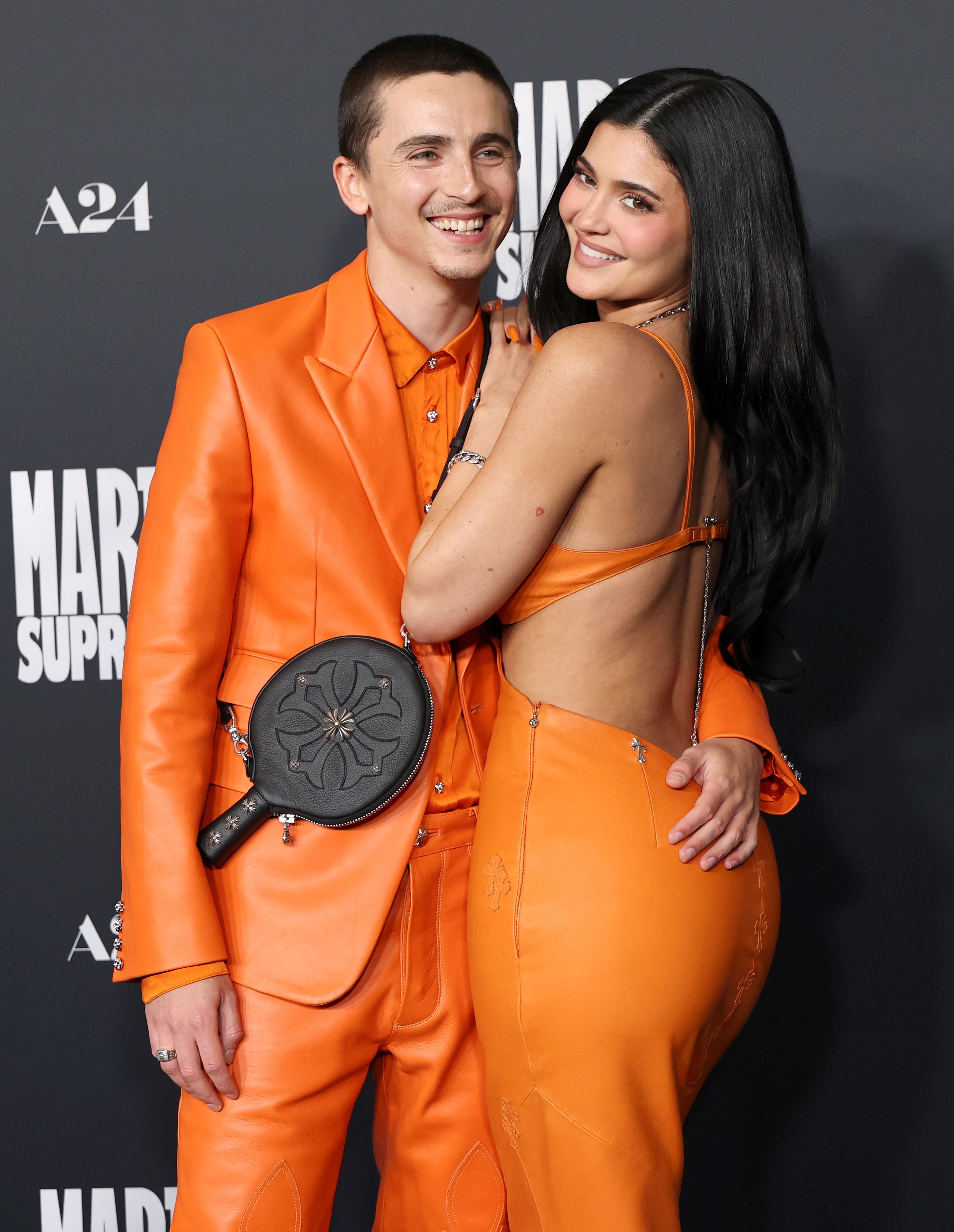 Two radical   connected  a reddish  carpet; 1  wears an orangish  suit, the different   a backless orangish  dress
