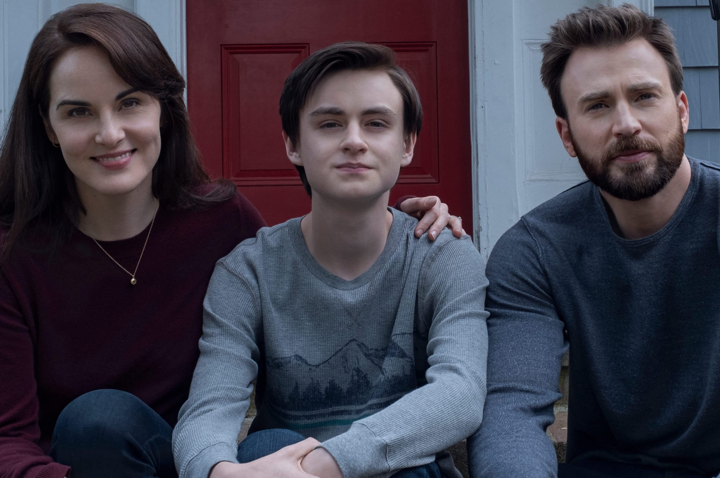 Three people sit together, smiling. The person in the middle wears a sweater with a mountain design, while the others wear simple long-sleeve tops