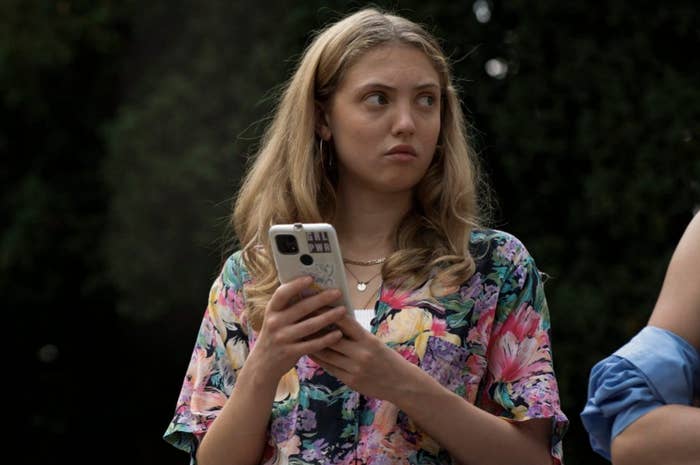 A woman with long hair holds a smartphone, wearing a floral patterned top, looking to the side with a thoughtful expression