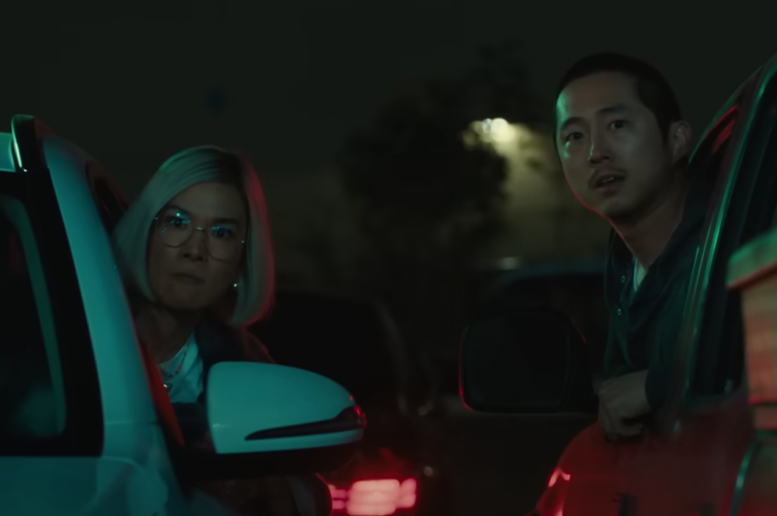 Two people look out of car doors in a dimly lit parking area, appearing surprised or concerned