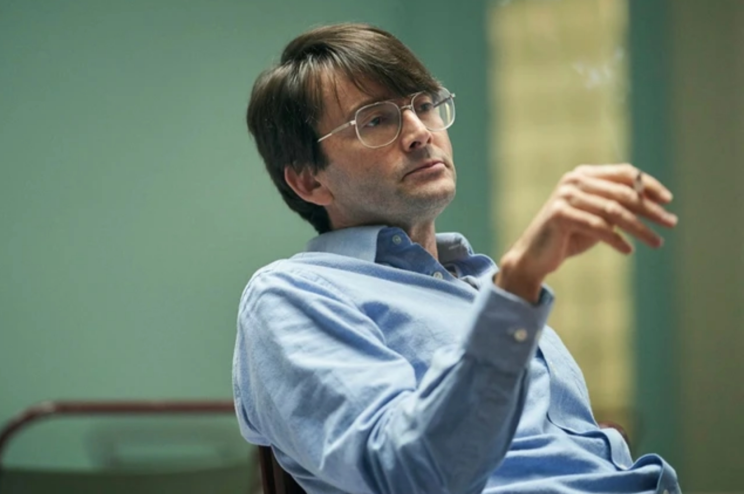 A person in glasses, wearing a button-down shirt, sits thoughtfully indoors, holding a cigarette