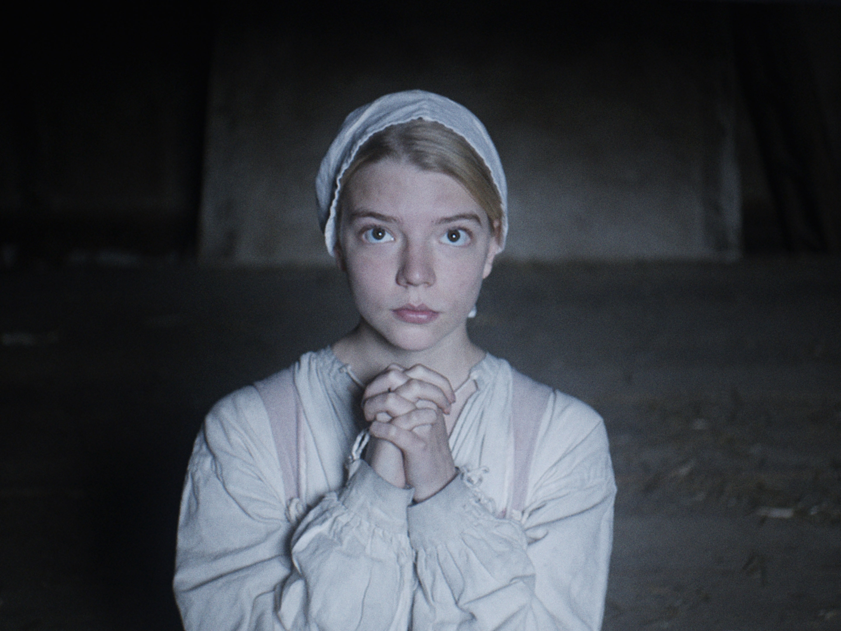 Taylor-Joy in historical attire with a bonnet and clasped hands, looking directly ahead in a dimly lit setting