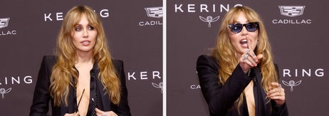 女性が黒いスーツとサングラスを着用し、赤いカーペットを歩いている。背景にはブランドロゴが見える。