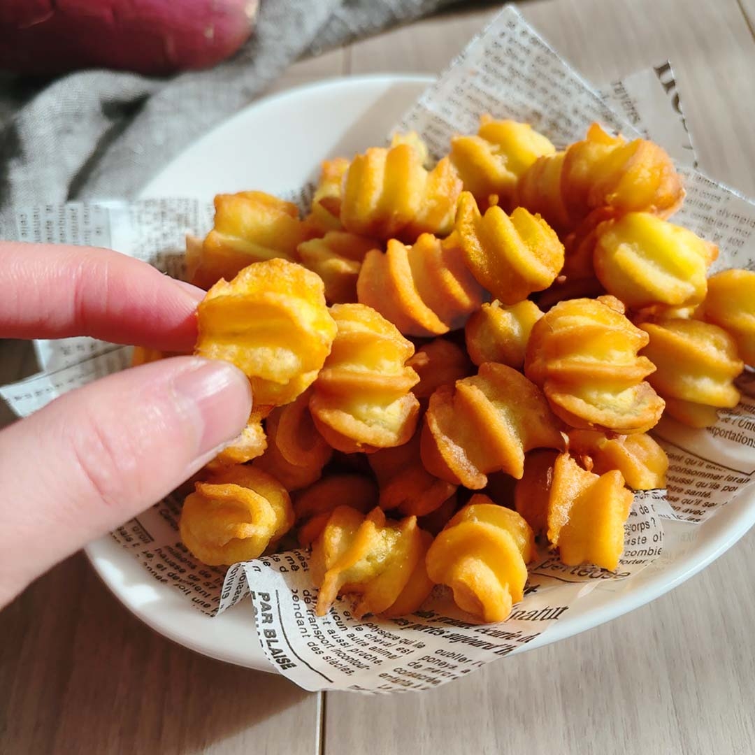 ボウルに入った揚げられたポテトスナックを手でつまんでいる様子。背景には新聞風の紙が敷かれている。