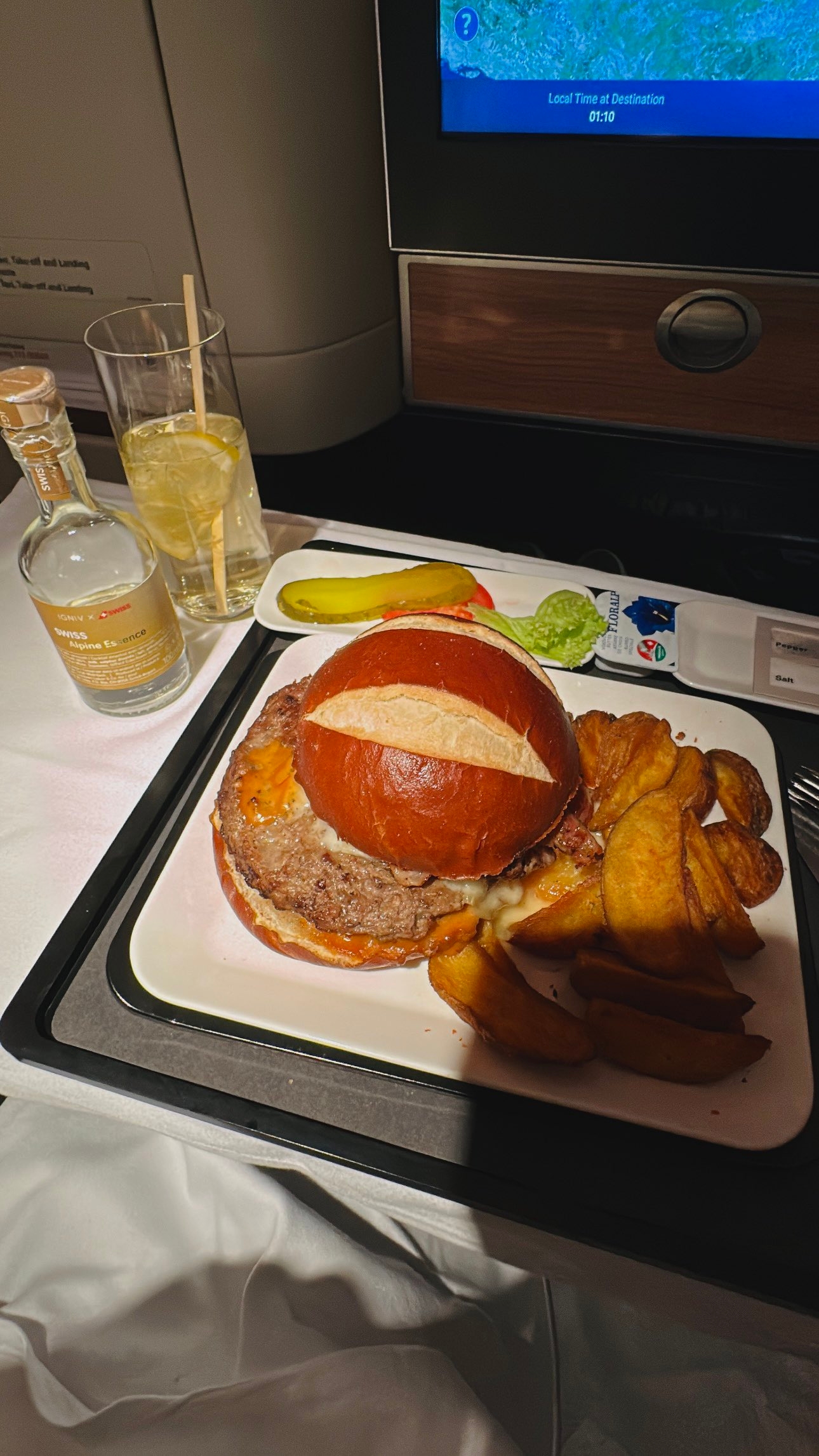 Gourmetburger med en kringlebolle, servert med tykke potetskiver, en sylteagurk og en flaske cocktail, på et flybrettbord