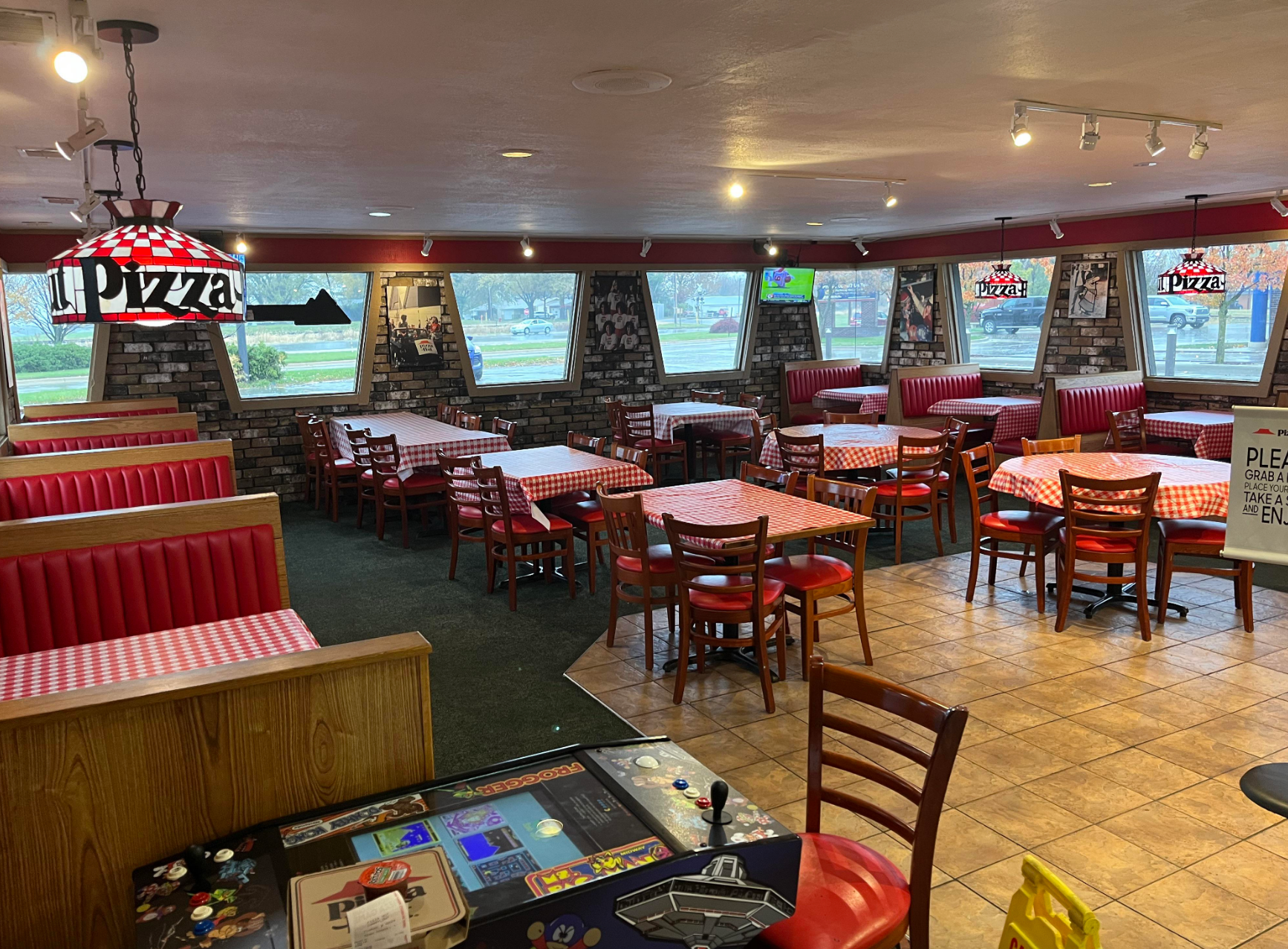 Empty pizzeria dining area with checkered tablecloths, wooden chairs, and booth seating, featuring a vintage arcade game table in the foreground
