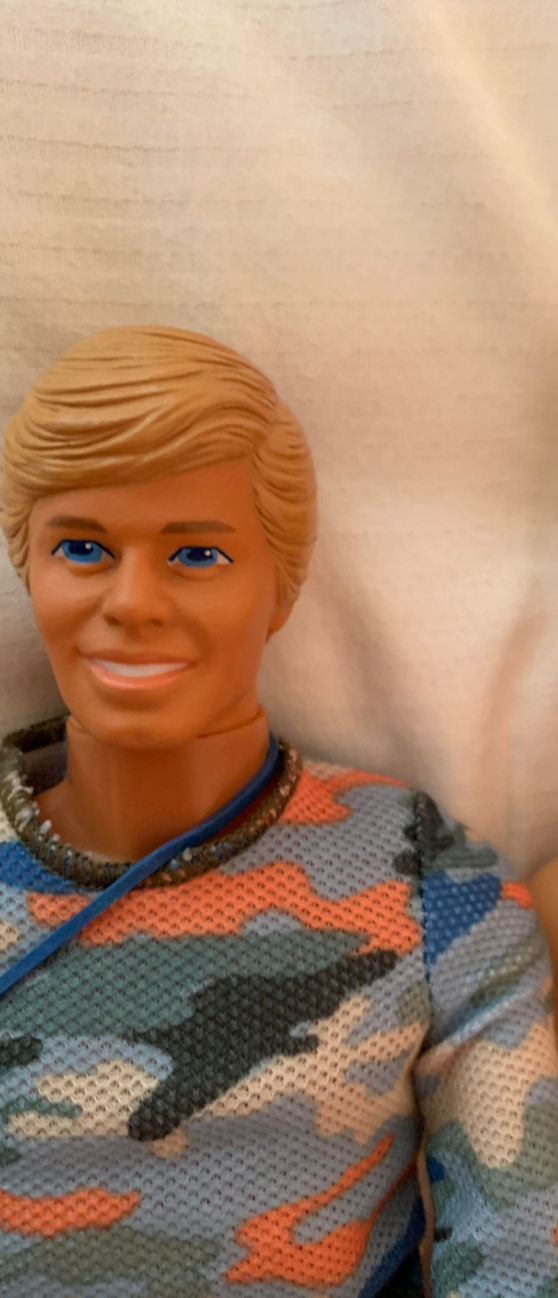 Plastic doll with short hair, wearing a knit sweater with a camouflage pattern, lying on a fabric surface