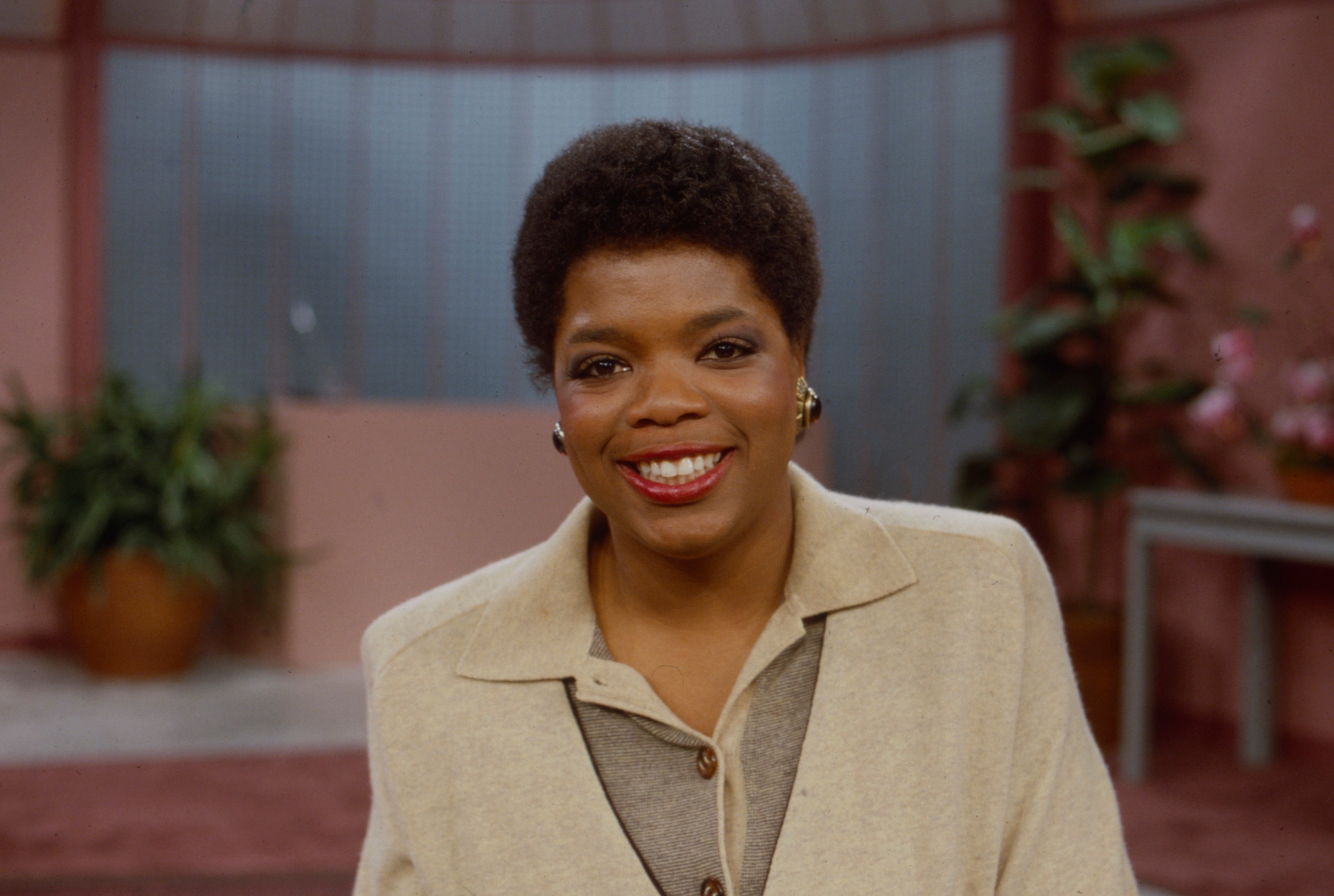 Person smiling successful  a workplace  setting, wearing a beige overgarment   implicit    a collared shirt. Background features potted plants and brushed  lighting