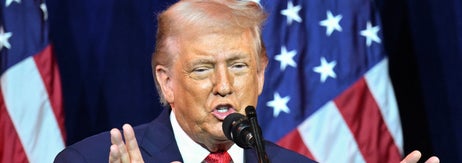 壇上でスーツの男性が演説し、後ろには星条旗が掲げられている。