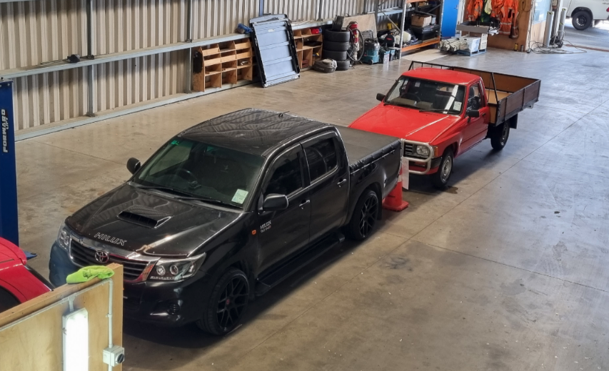 Two pickup trucks are parked in a workshop, with an orange cone placed between them
