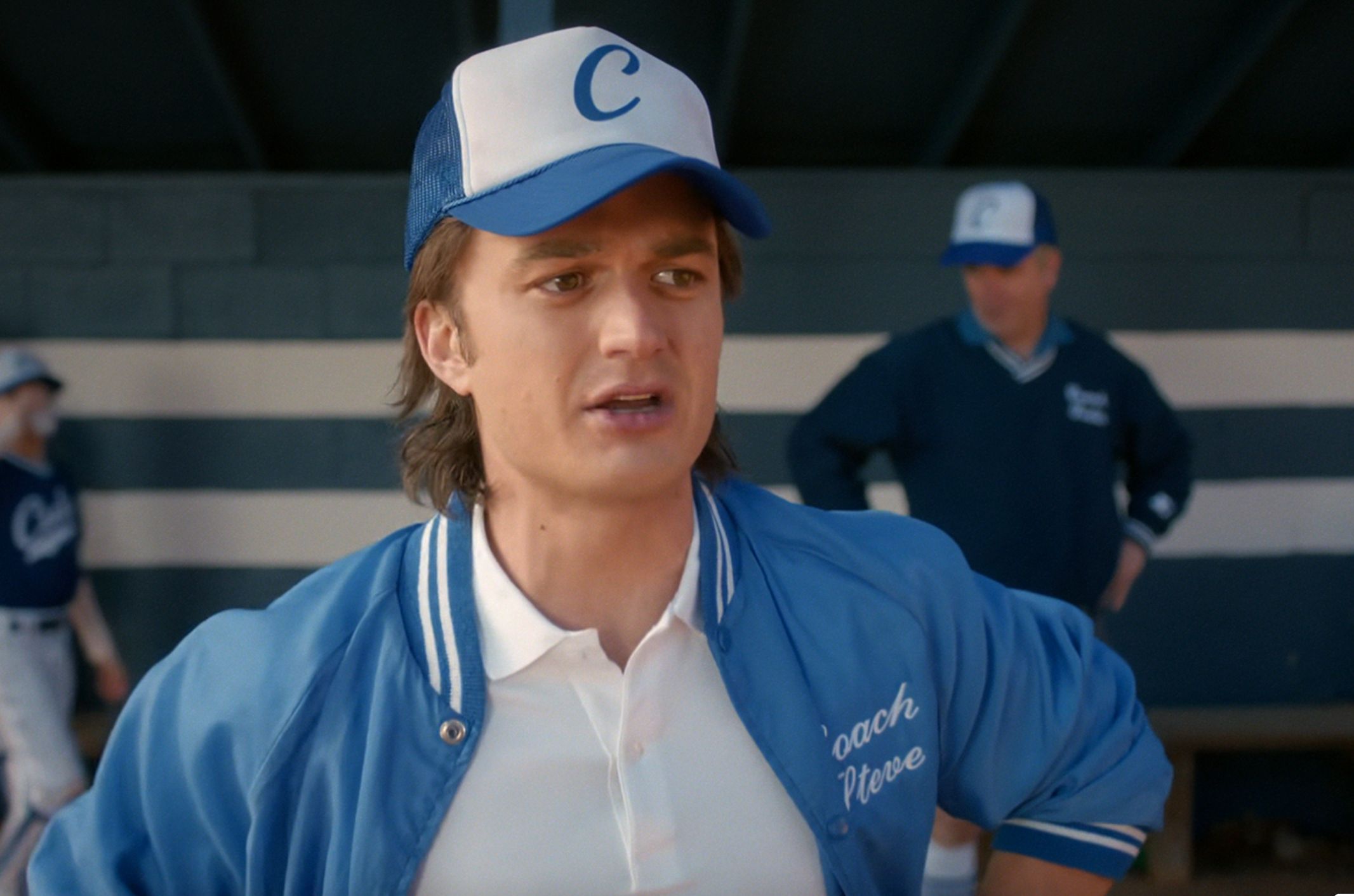 Person in a baseball uniform and cap, wearing a jacket with "Coach Steve" embroidered, standing on a baseball field. Players in the background
