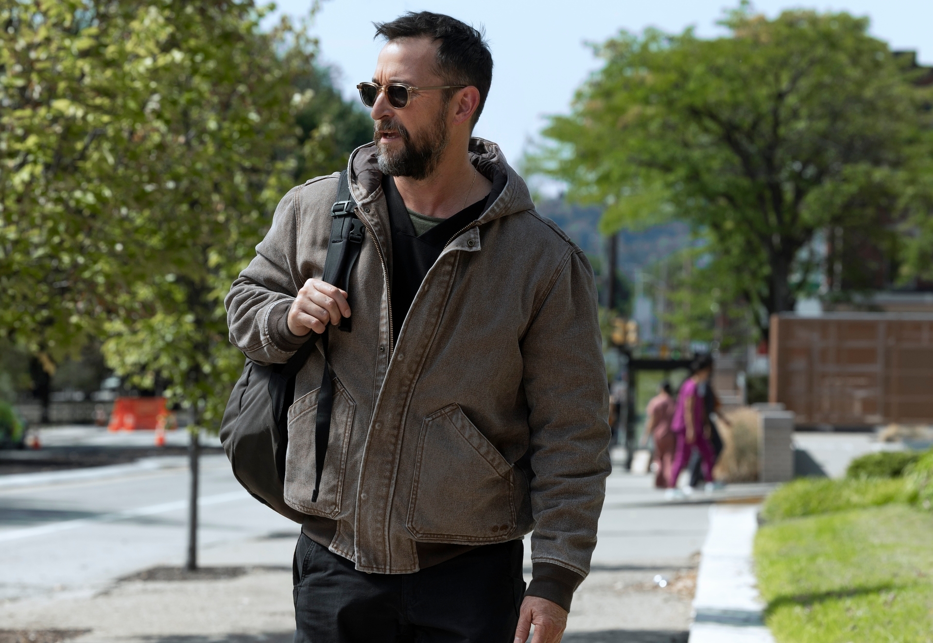 A idiosyncratic   walks outdoors, wearing a casual overgarment   and sunglasses, carrying a backpack implicit    1  shoulder, with trees and buildings successful  the background
