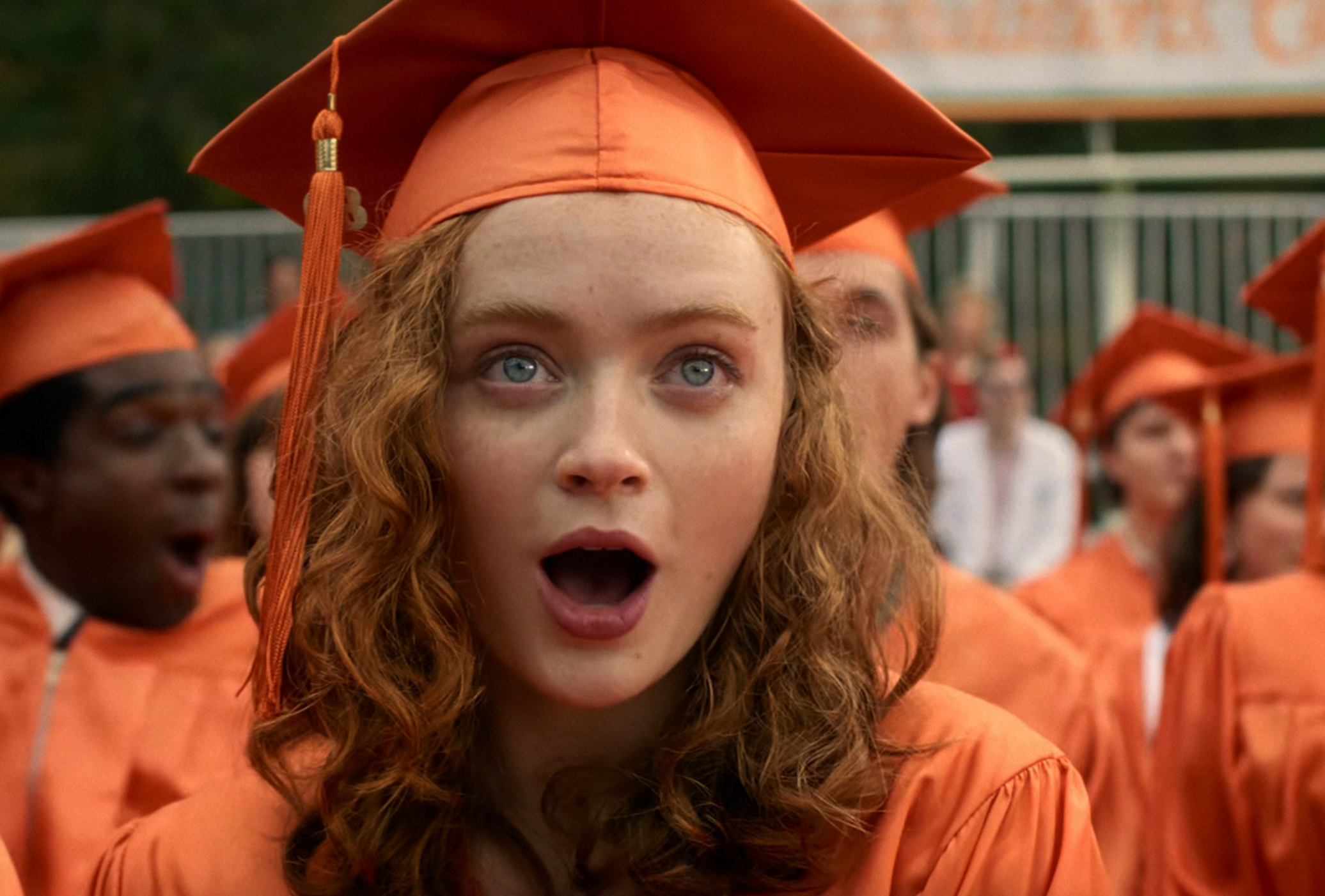 A young graduate in an orange cap and gown stands among classmates, looking surprised during a commencement ceremony