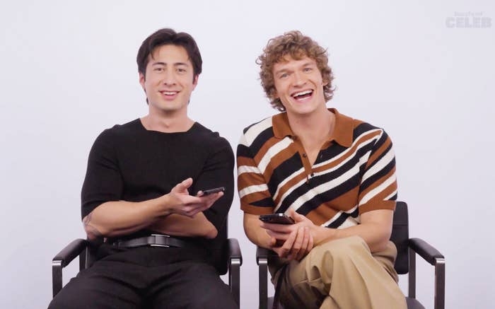 Two radical   sitting broadside  by side, smiling and holding phones. One wears a coagulated  outfit, the different   a patterned garment  with stripes