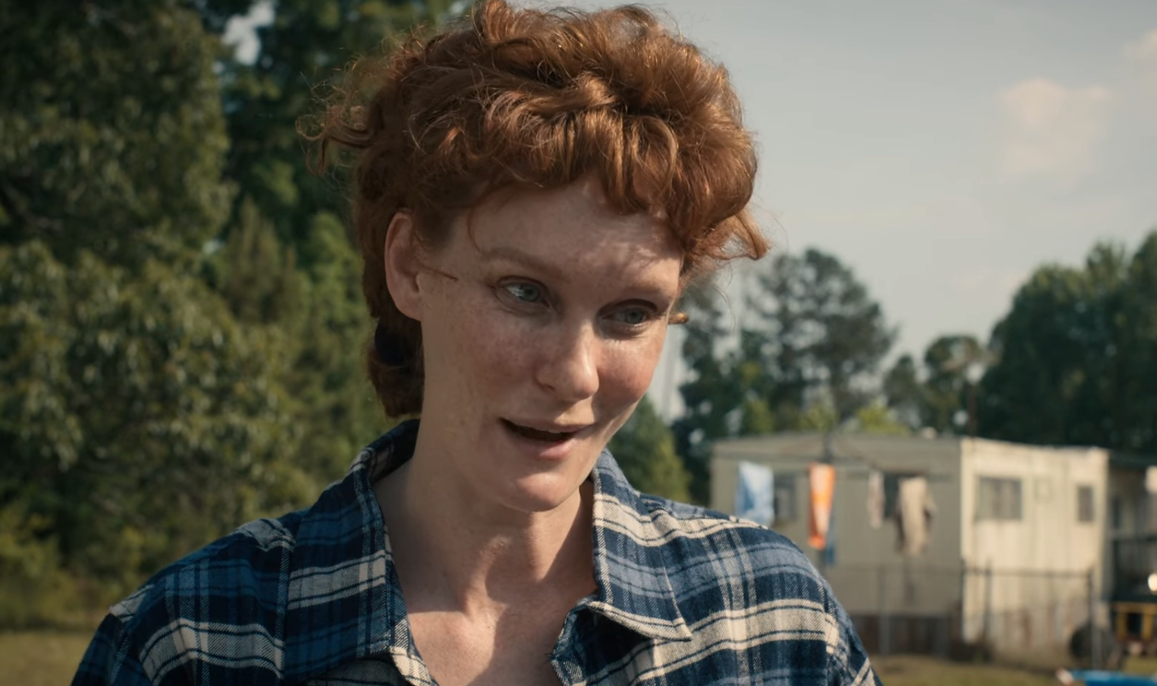 Person outdoors wearing a plaid shirt, standing in front of a trailer with trees in the background