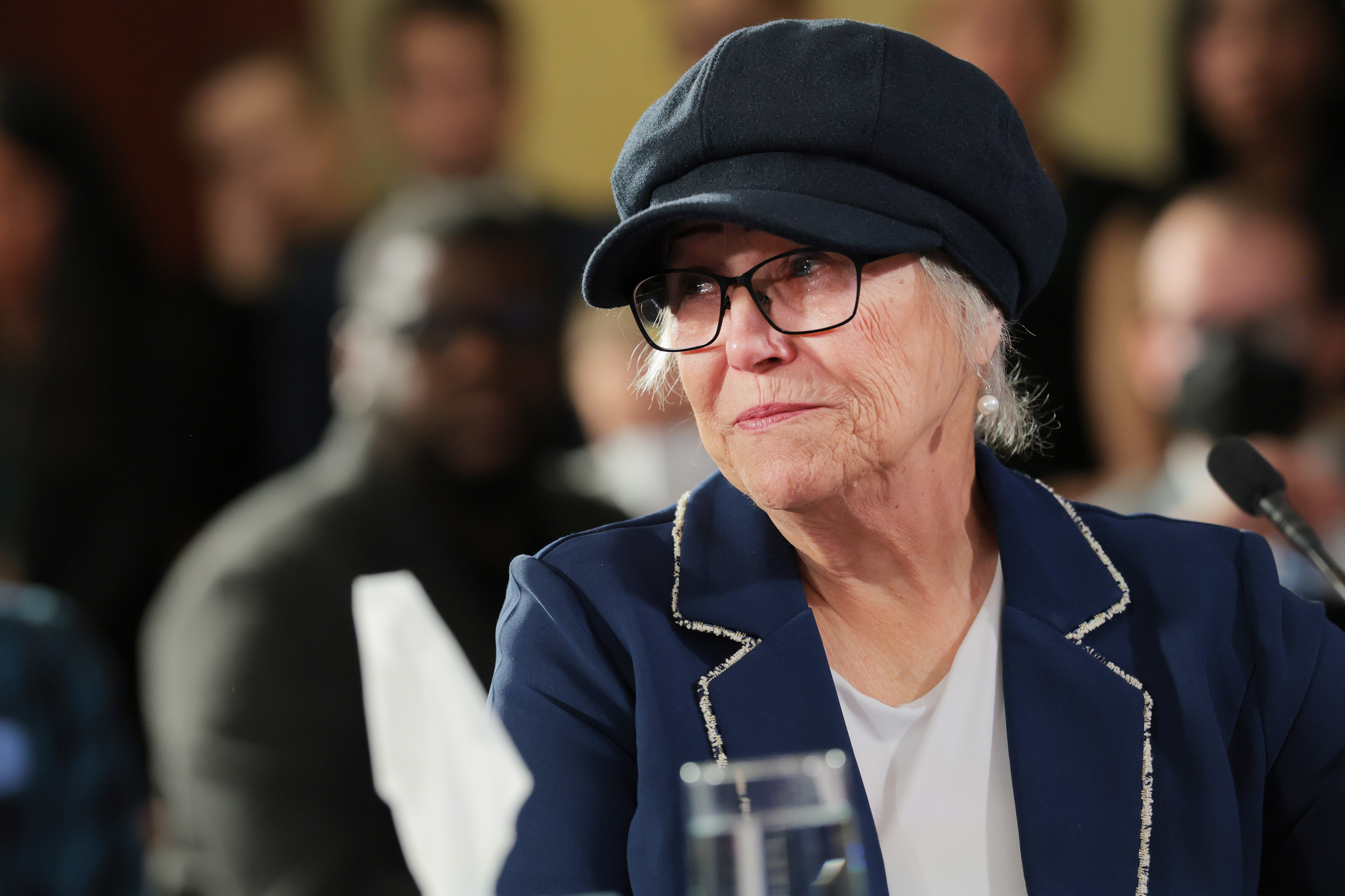 Person i blazer og caps, snakker ved en mikrofon, i en formell setting med et publikum i bakgrunnen