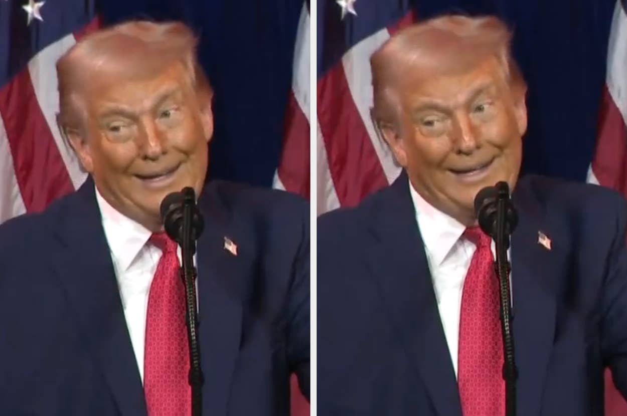 Man at podium with flags in background, hair appears humorously distorted as if blown by wind