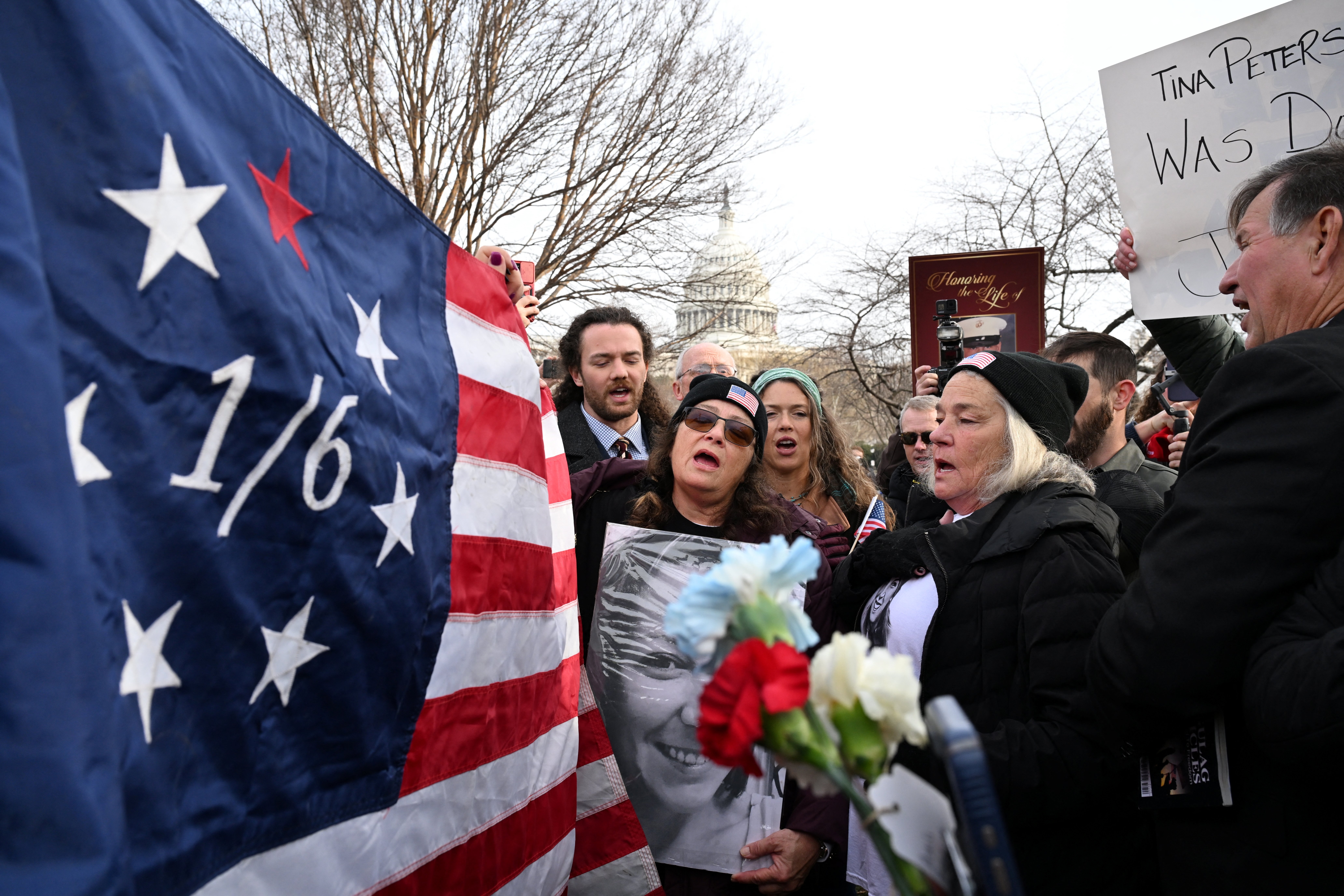 Gruppe som holder et flagg med "1/6," blomster og skilt foran US Capitol under en protest