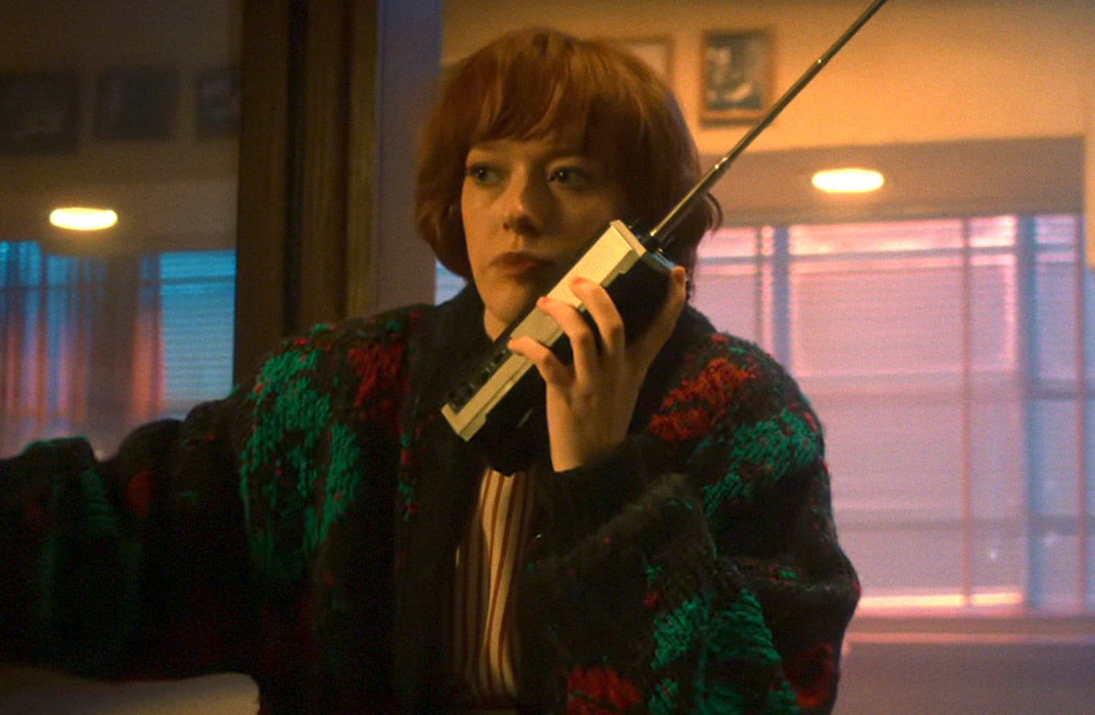 Person in a cozy sweater holds a vintage portable phone, appearing concerned while looking out a window